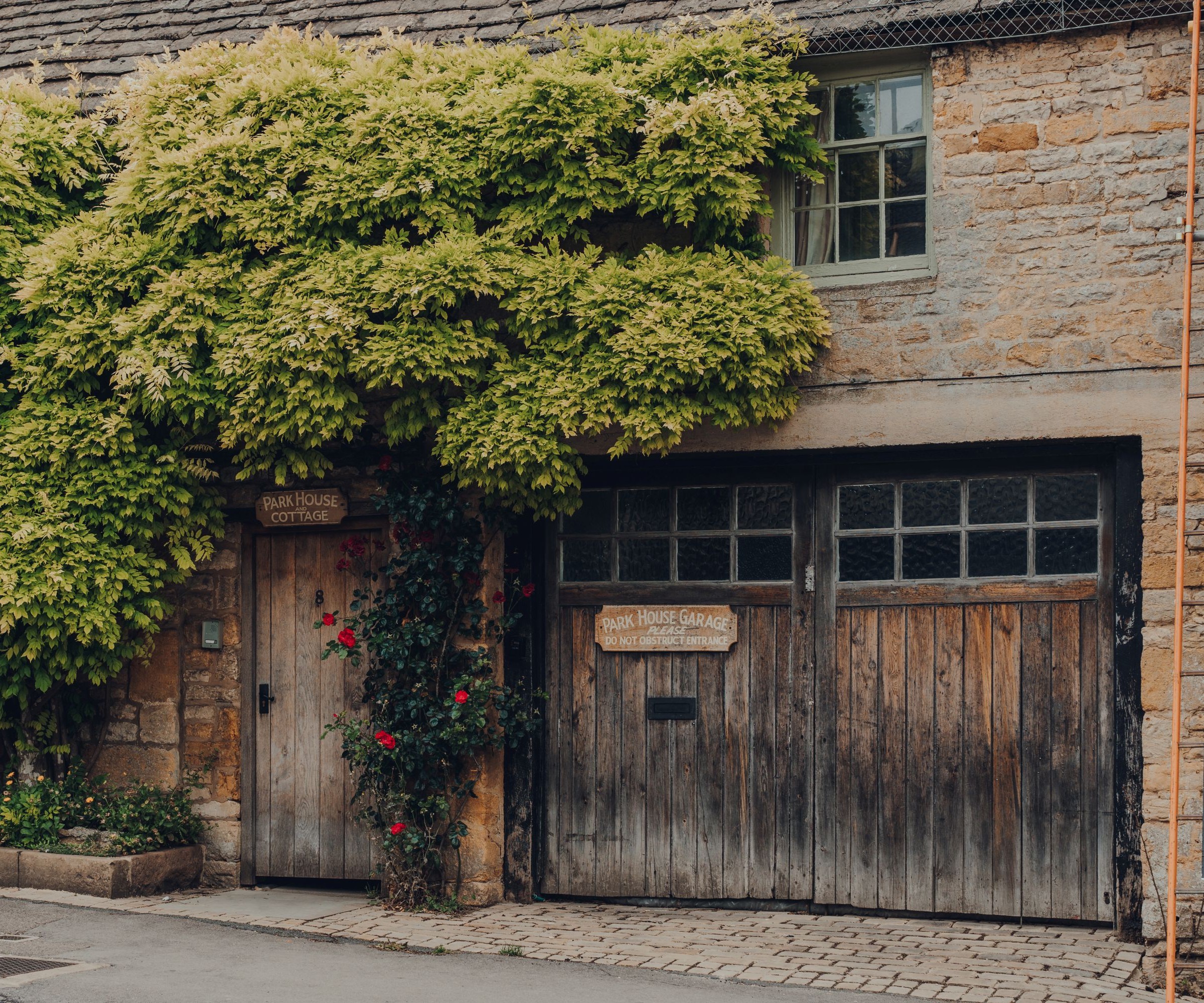 An historic looking garage with an old stained wooden garage door with windows and a living area on the second storey above it. A large plant climbing the brickwork is also in the image