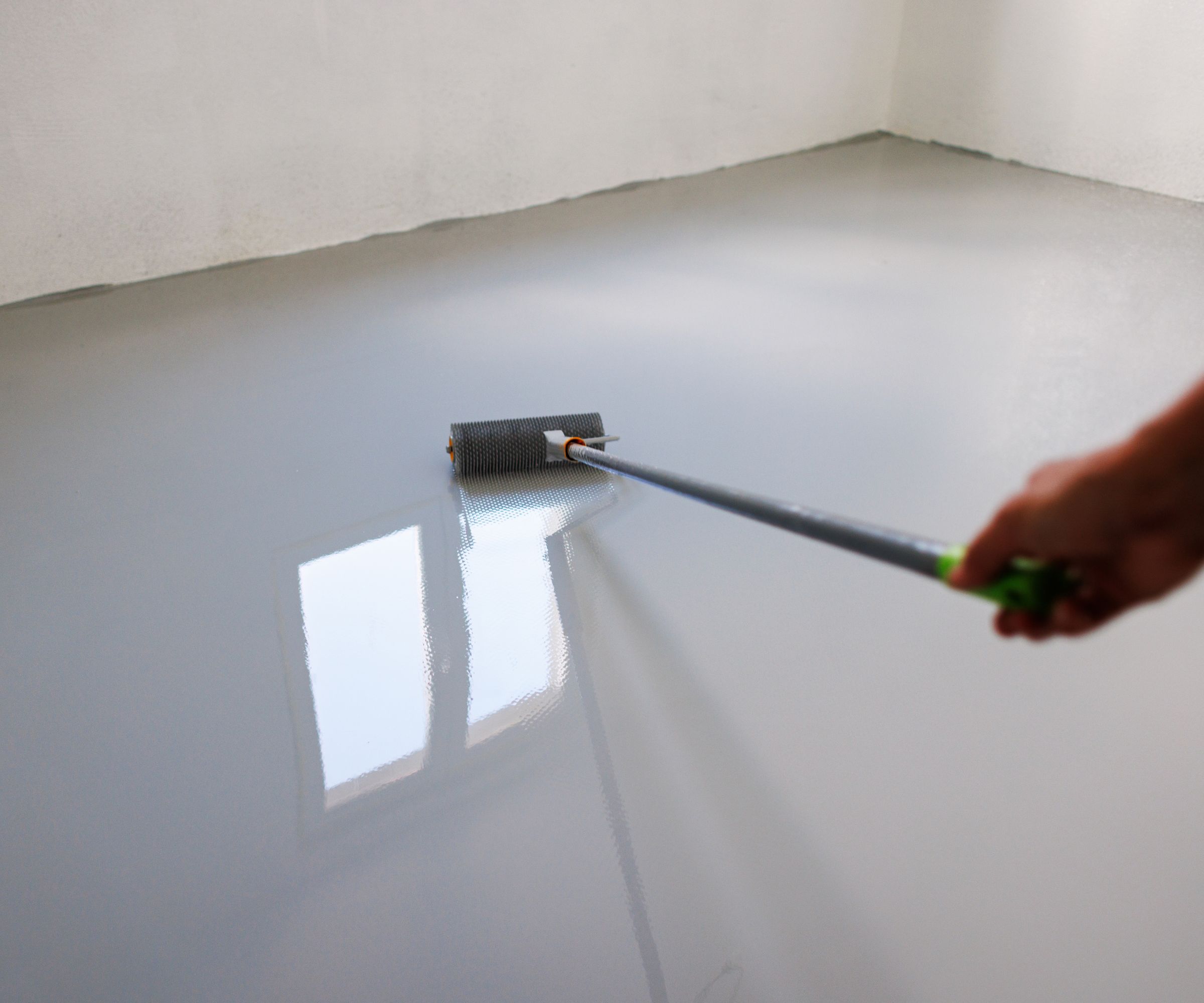 A close-up of a hand using a spiked roller to apply epoxy resin coating on a newly leveled floor. The reflective surface highlights the professional finishing process in a bright, natural-light environment. Ideal for illustrating industrial flooring, home renovation, or construction projects.