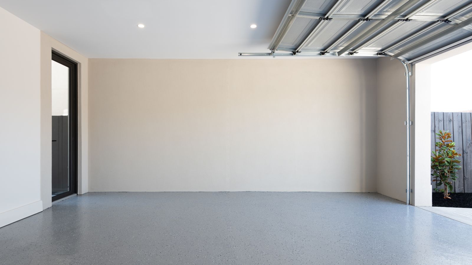 A clean, modern garage with smooth, speckled grey flooring and cream walls. The ceiling is white with recessed spotlights, and the metal garage door is partially open. A black-framed glass door leads to another part of the home, and a small tree is visible just outside next to a timber fence.