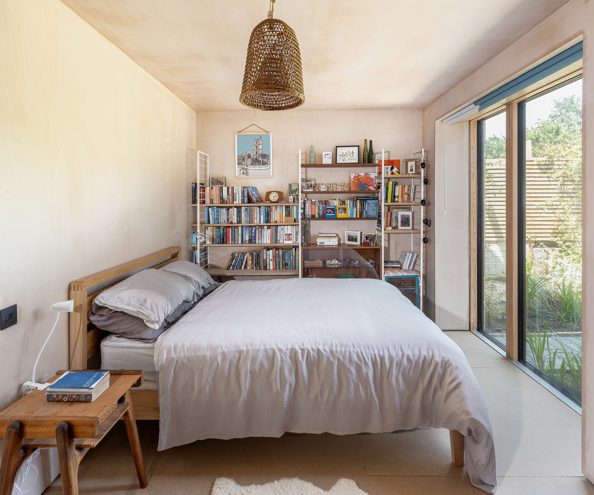 A ground floor bedroom in a self build home. There is a double bed and a set of shelves. The walls and ceiling are in a plaster. The bed us facing a glass sliding door which leads outside