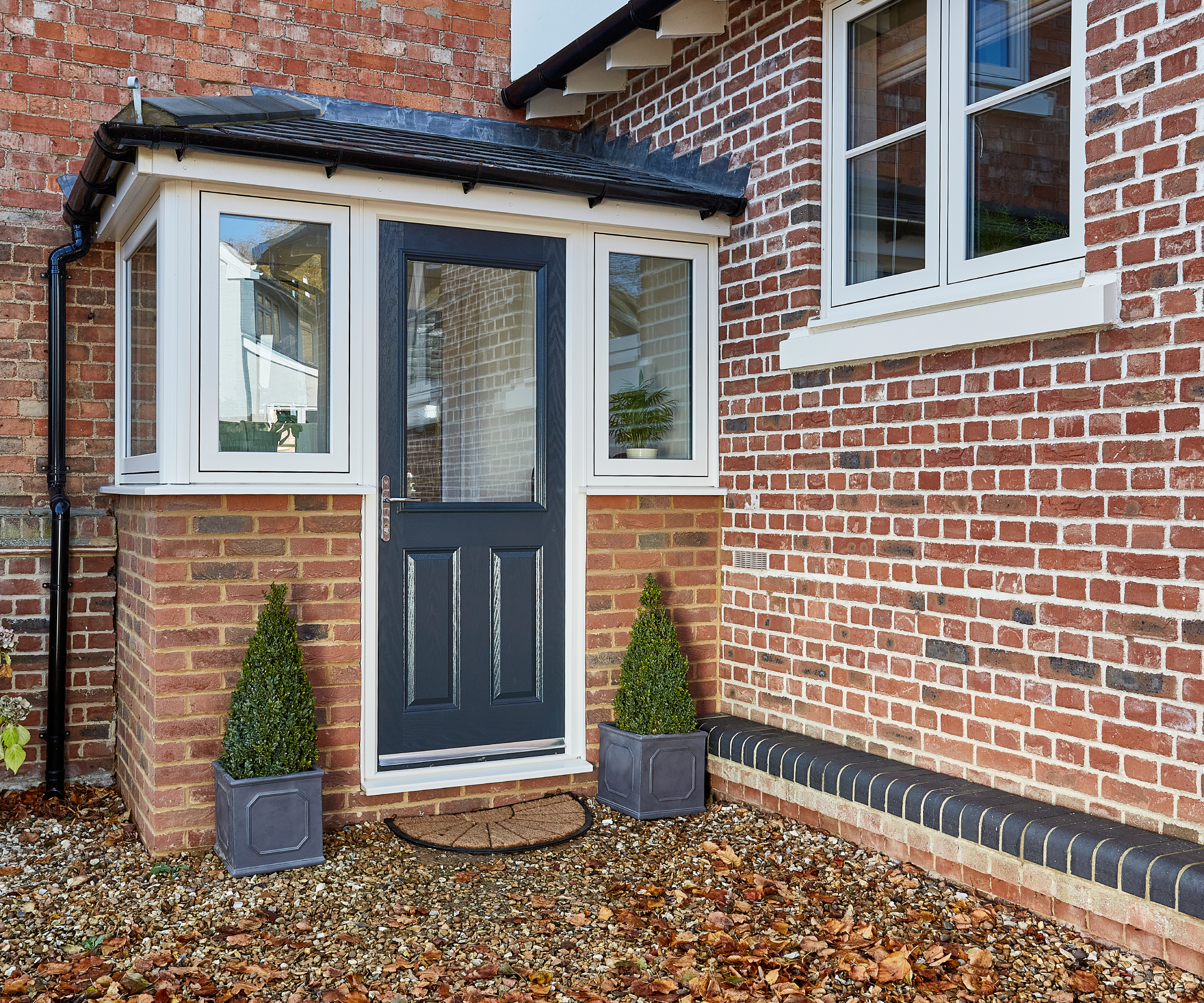 small enclosed brick porch with cream windows, grey door and slate tiled roof next to brick house and brick extension