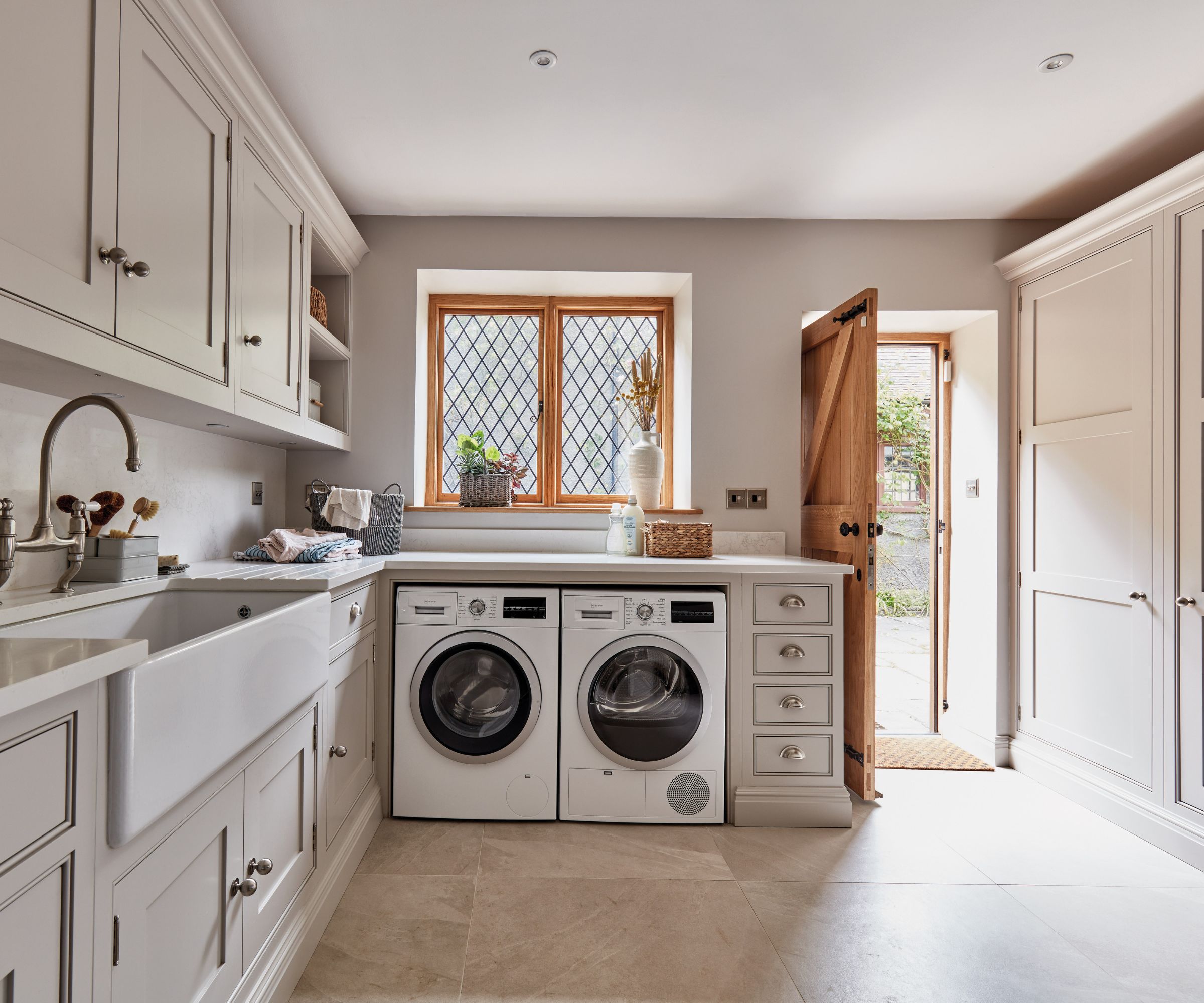 A beautifully designed utility room featuring classic shaker-style cabinetry in a soft, neutral tone. Twin front-loading washer and dryer units sit beneath a bright window with leaded panes, flanked by built-in drawers and storage.
