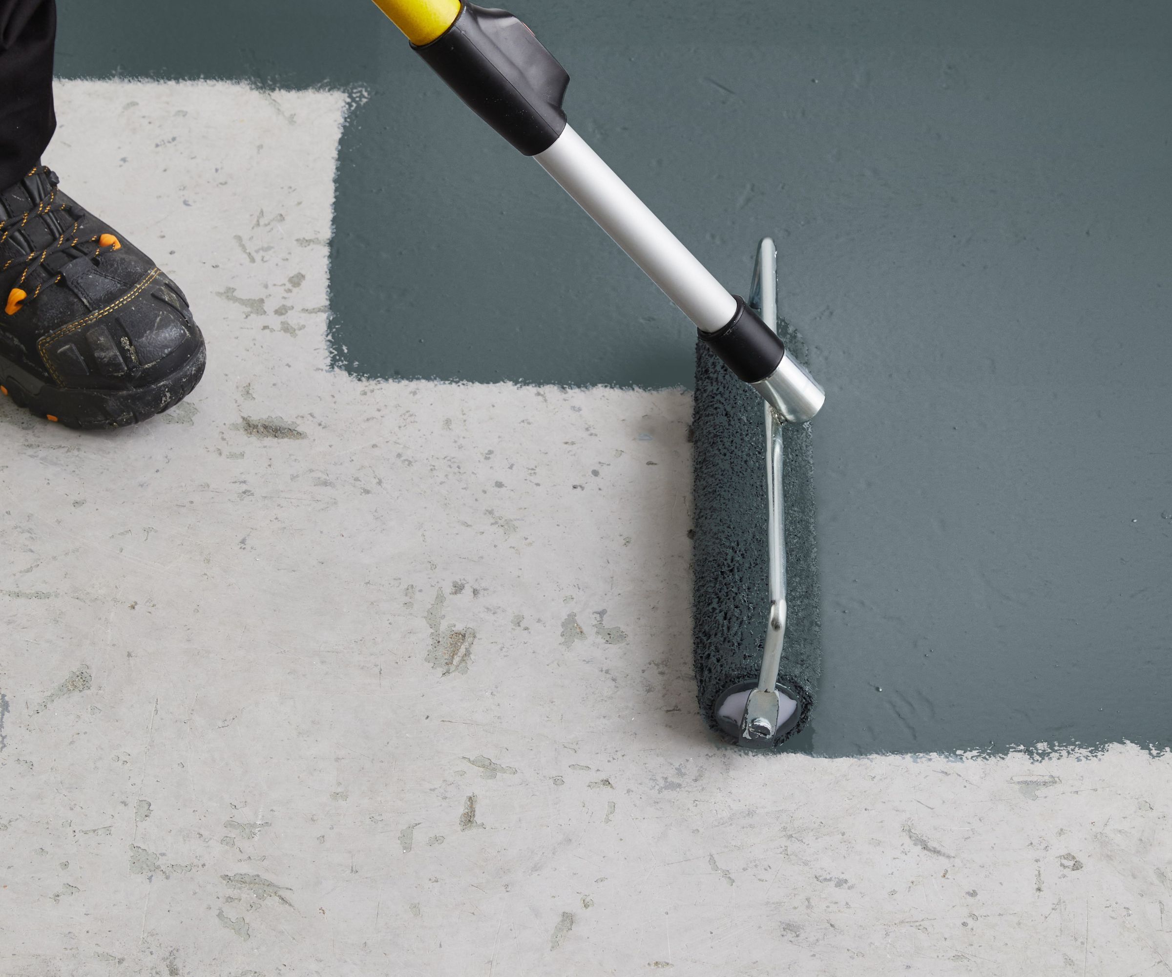 A close-up view of a person painting a concrete garage floor with dark grey epoxy paint using a roller attached to a long handle. The roller is mid-stroke, showing a clear contrast between the raw, unfinished concrete and the freshly coated surface