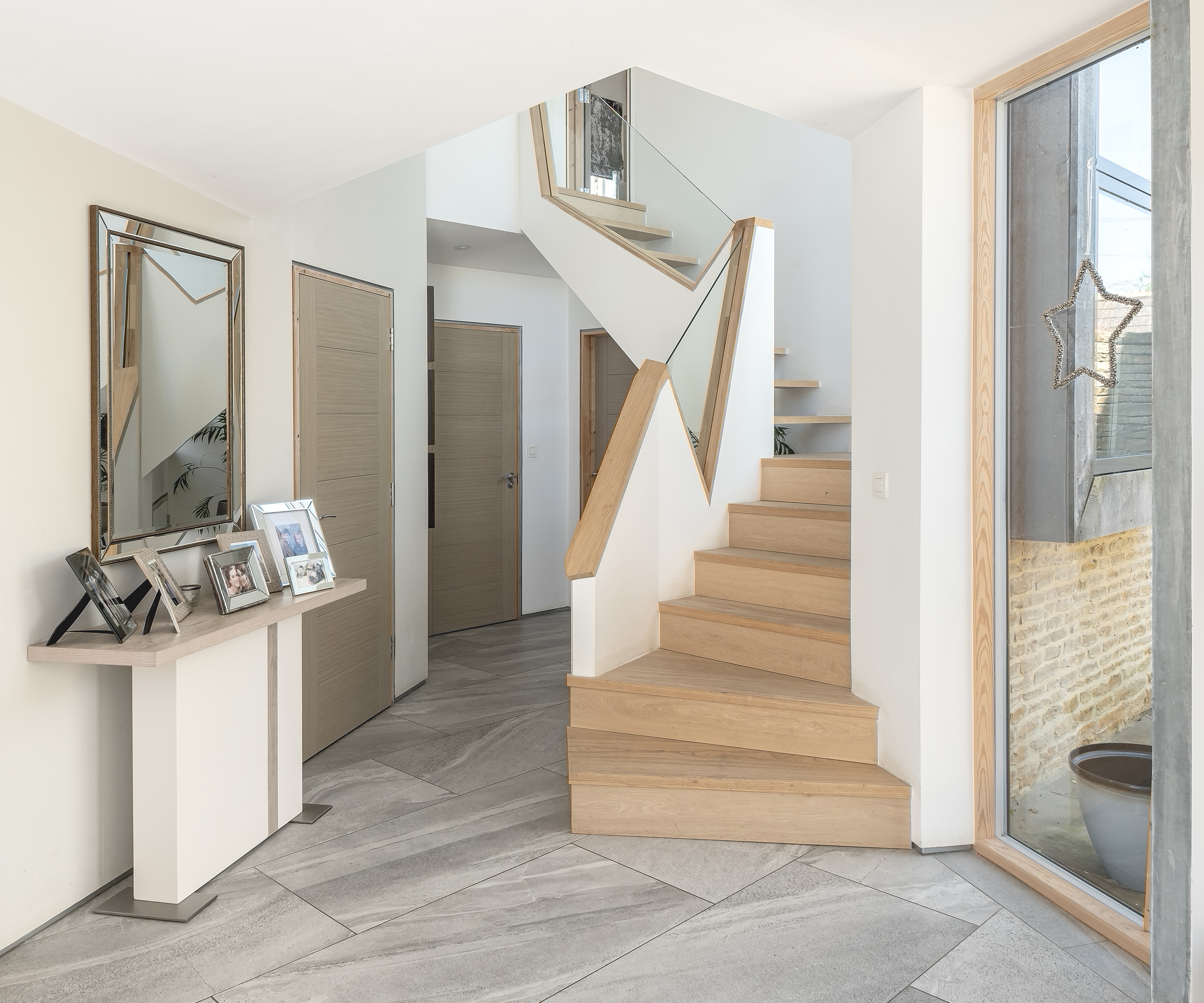 A contemporary hallway in a self-build home. The staircase has an abstract design with wood cutouts and glass. The flooring is plain grey but arranged at an angle