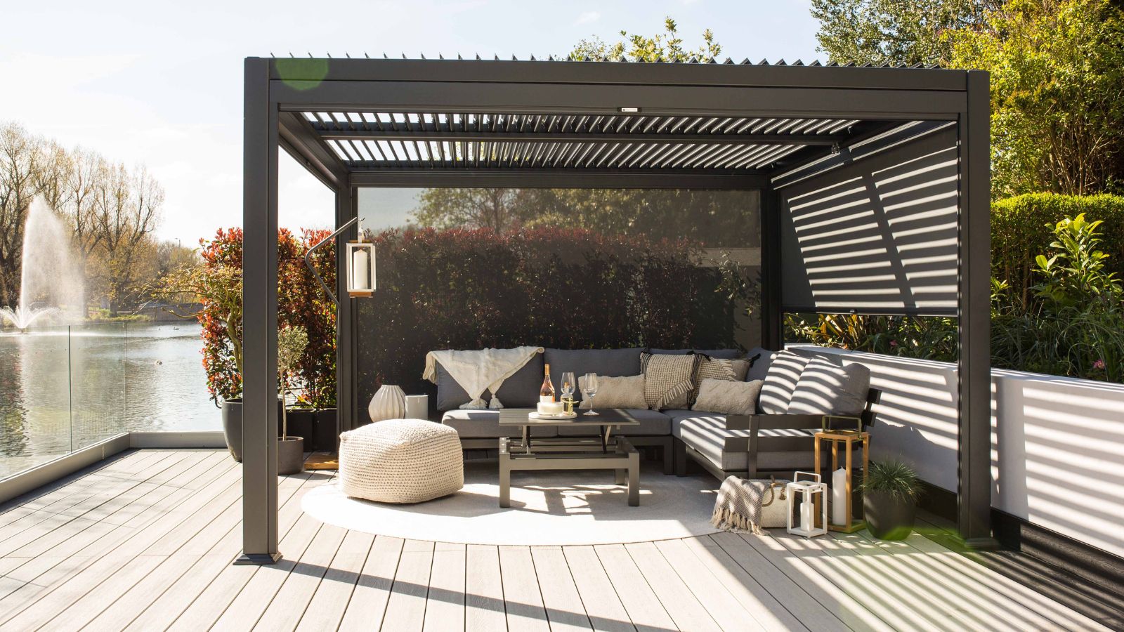 A modern pergola with a dark grey aluminium frame and a louvred roof stands on a light wood deck. Inside, a grey L-shaped sofa with textured cushions and throws surrounds a low coffee table set with drinks. A cream knitted pouffe, lanterns, and potted plants add warmth.