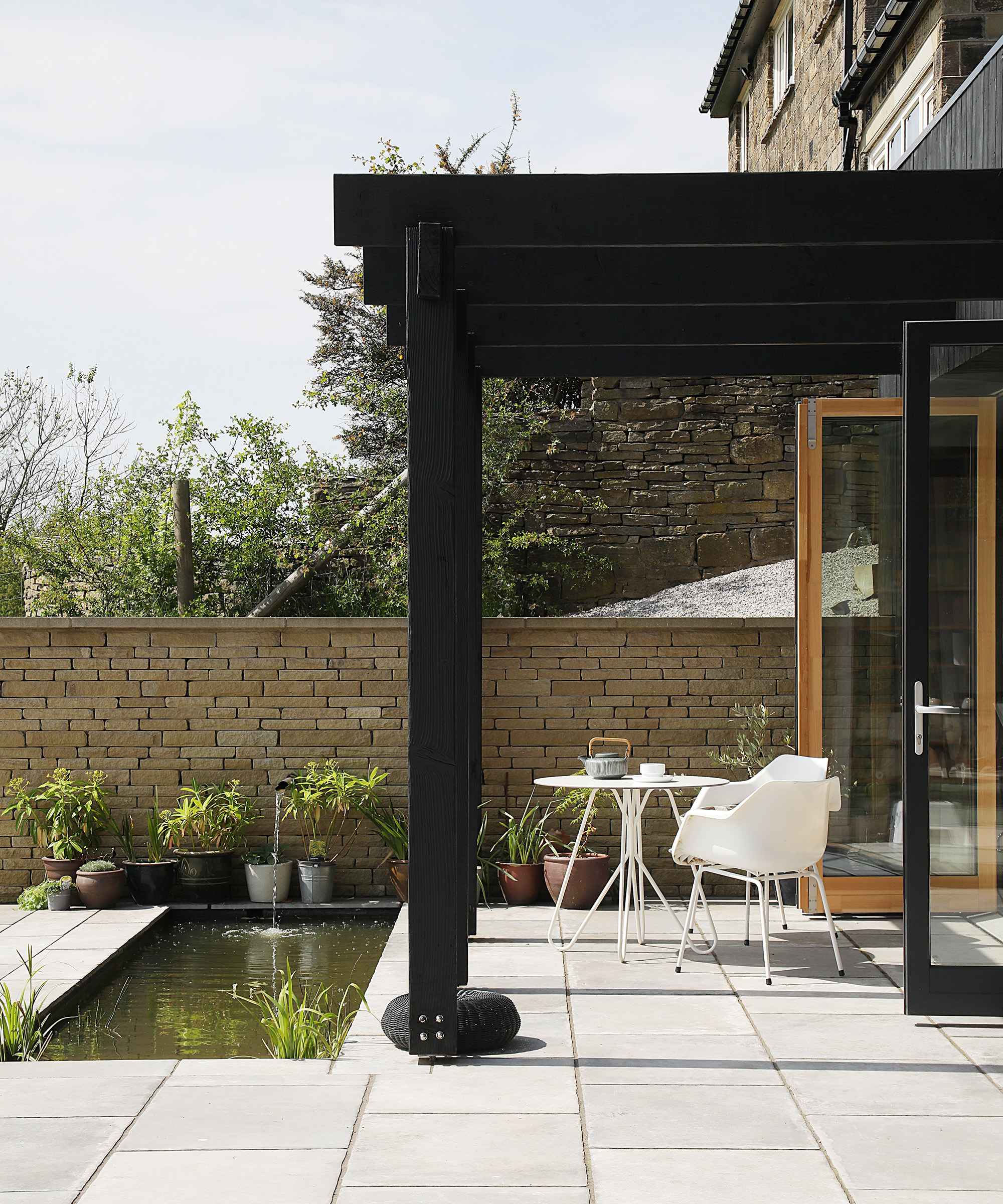 A stone patio area with white outdoor table and chairs set. There is a rectangular pond built into the paving and a black timber frame structure