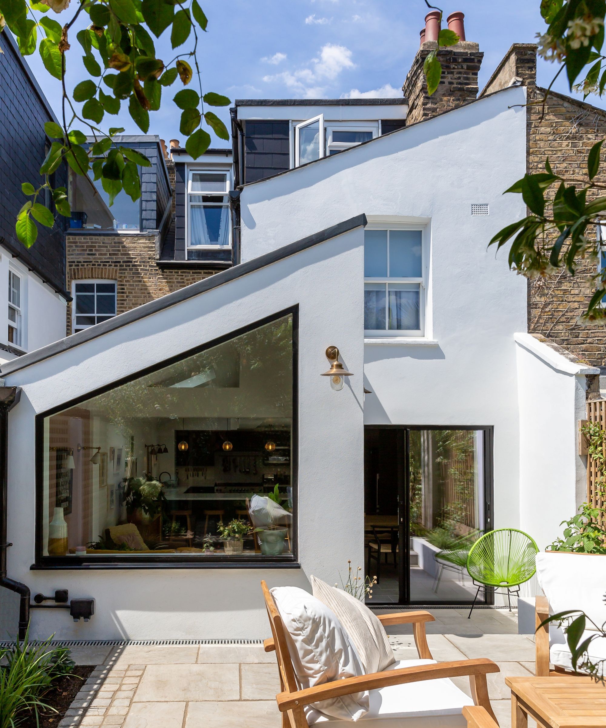 A white rendered extension with a sloped roof and large unusual shaped window and the original brick and timber clad home seen behind it