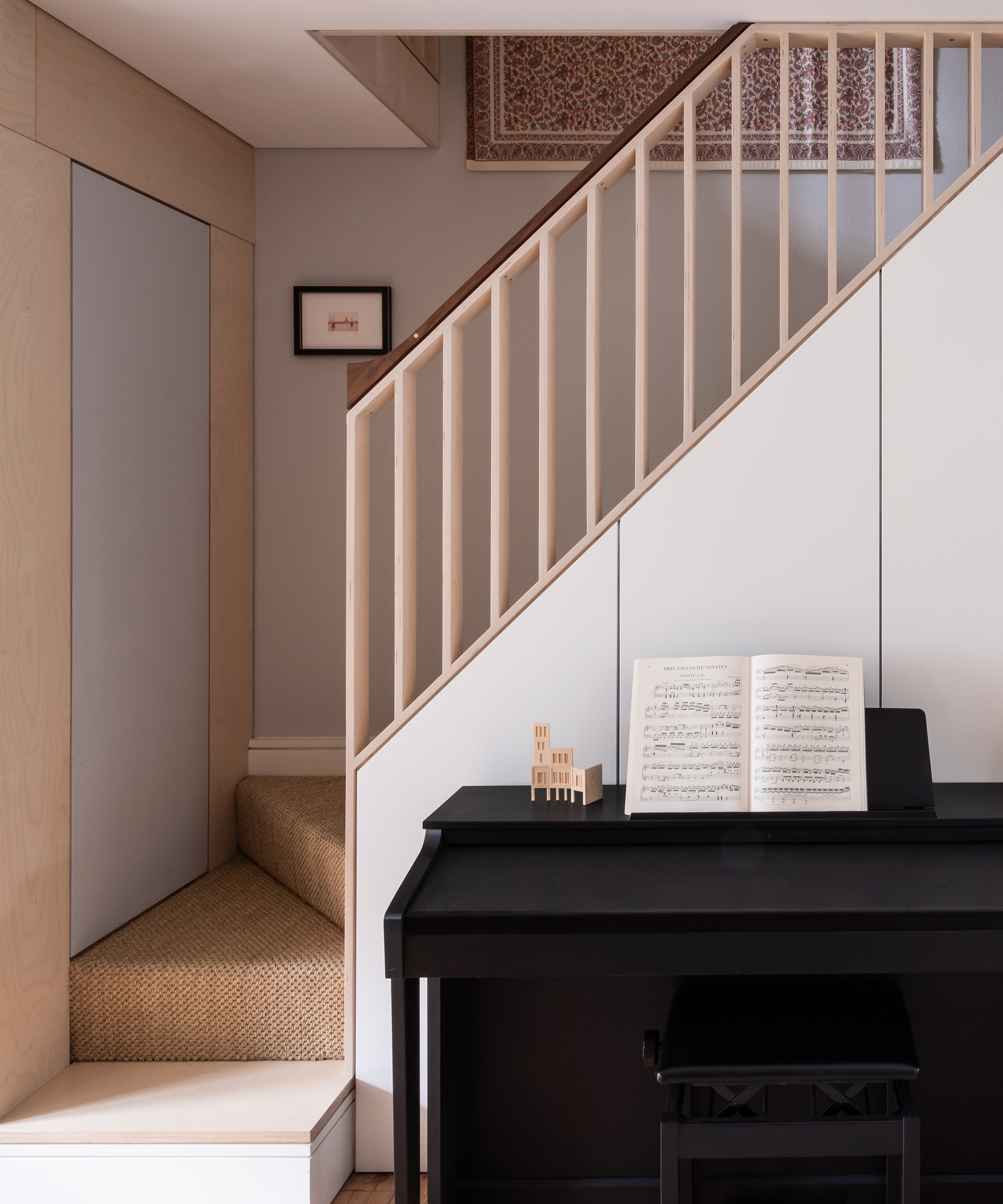 A piano sits in front of a small staircase in a Victorian terrace house with plywood bannister rails