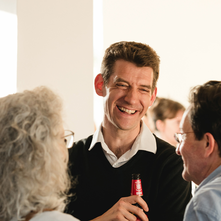 male with short brown hair wearing black jumper and white shirt in social setting talking to another male and female