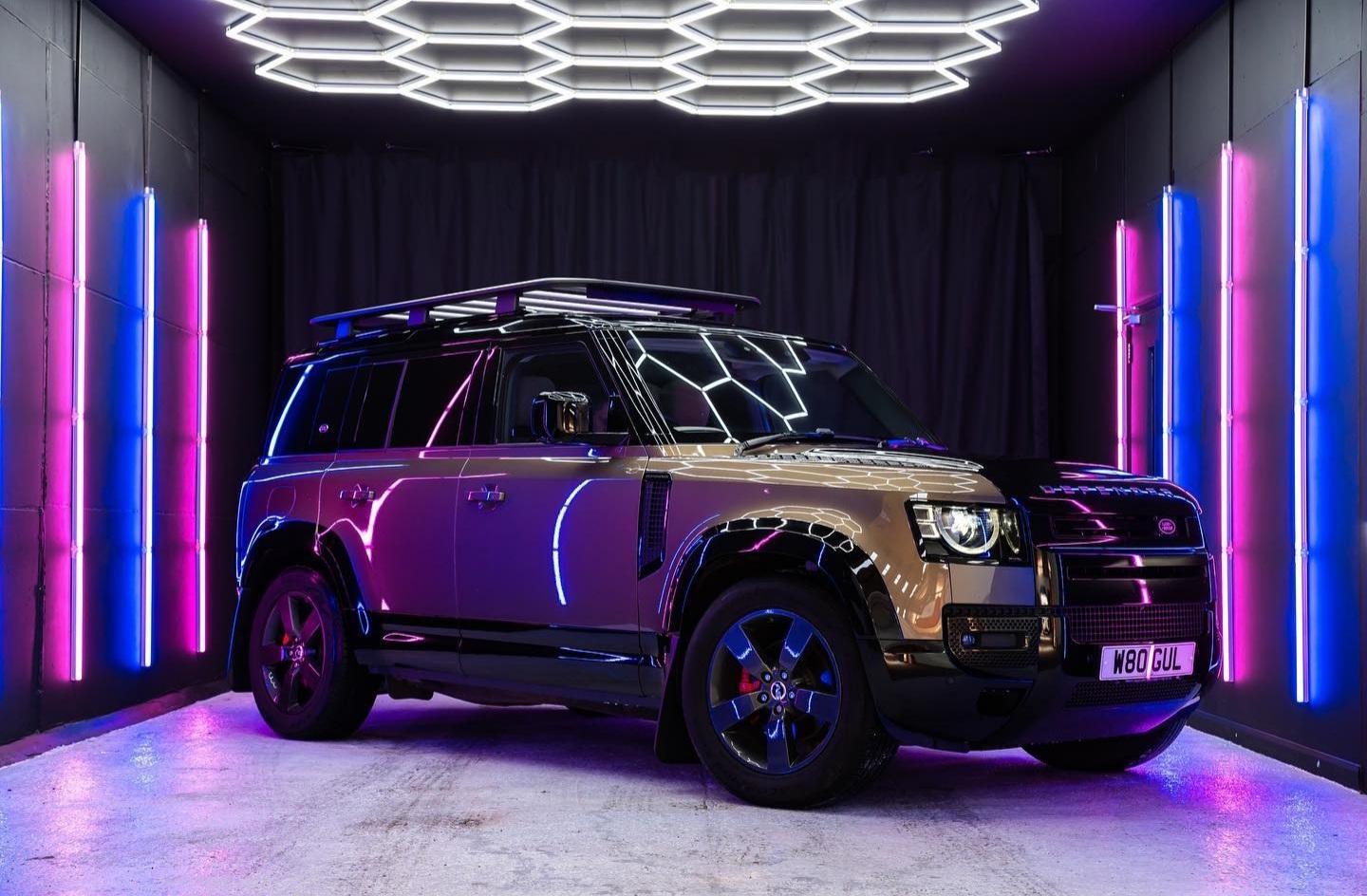 A black and gold Land Rover parked inside a garage illuminated with dramatic neon lighting. The ceiling features white hexagon-shaped LED lights, while the walls glow with vertical strips of pink, purple, and blue lights, creating a futuristic showroom effect.
