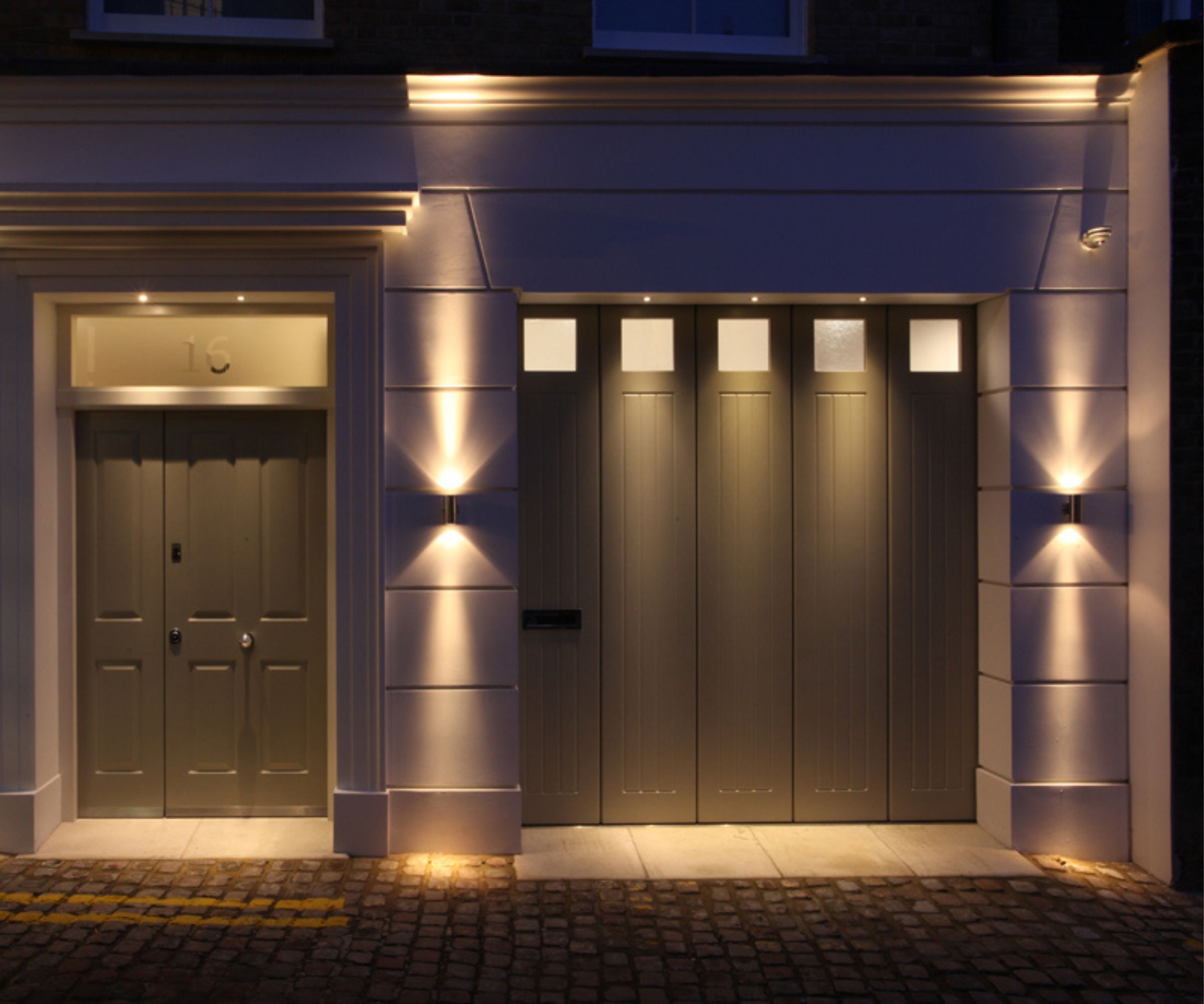 A townhouse exterior at night with a front door and garage door illuminated by warm outdoor lighting. Small recessed lights highlight the doorway and garage.