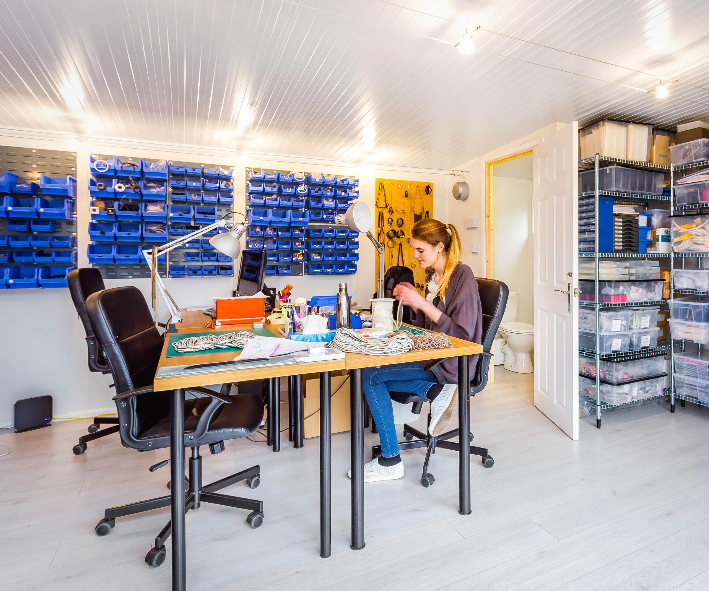 A garage workshop with desks and office chairs. Blue wall-mounted storage bins and clear shelving units line the walls. A woman sits at a desk working with craft materials.
