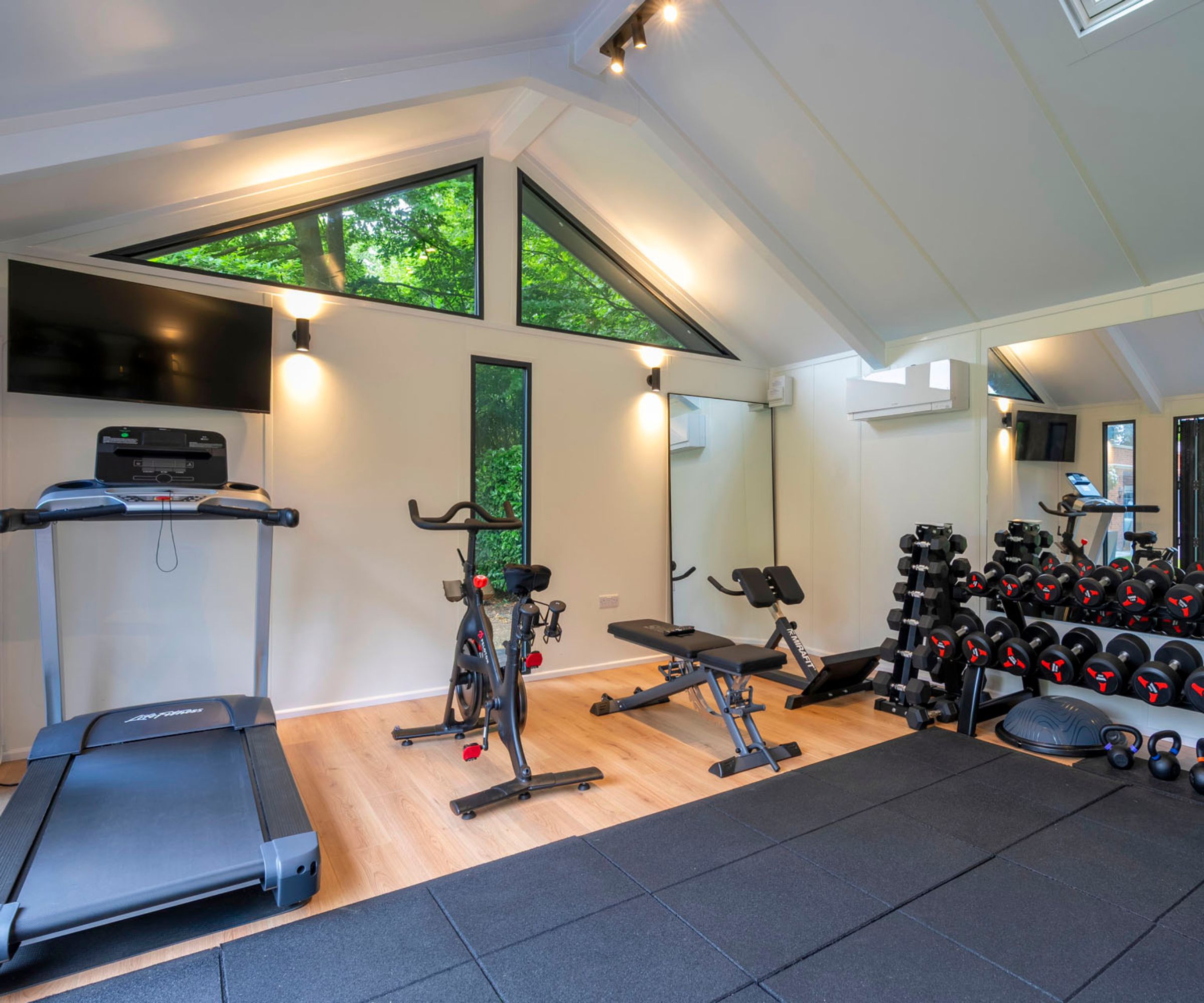 A bright garage converted into a home gym, with treadmills, bikes, and weights. Spotlights and natural light from triangular windows illuminate the room.