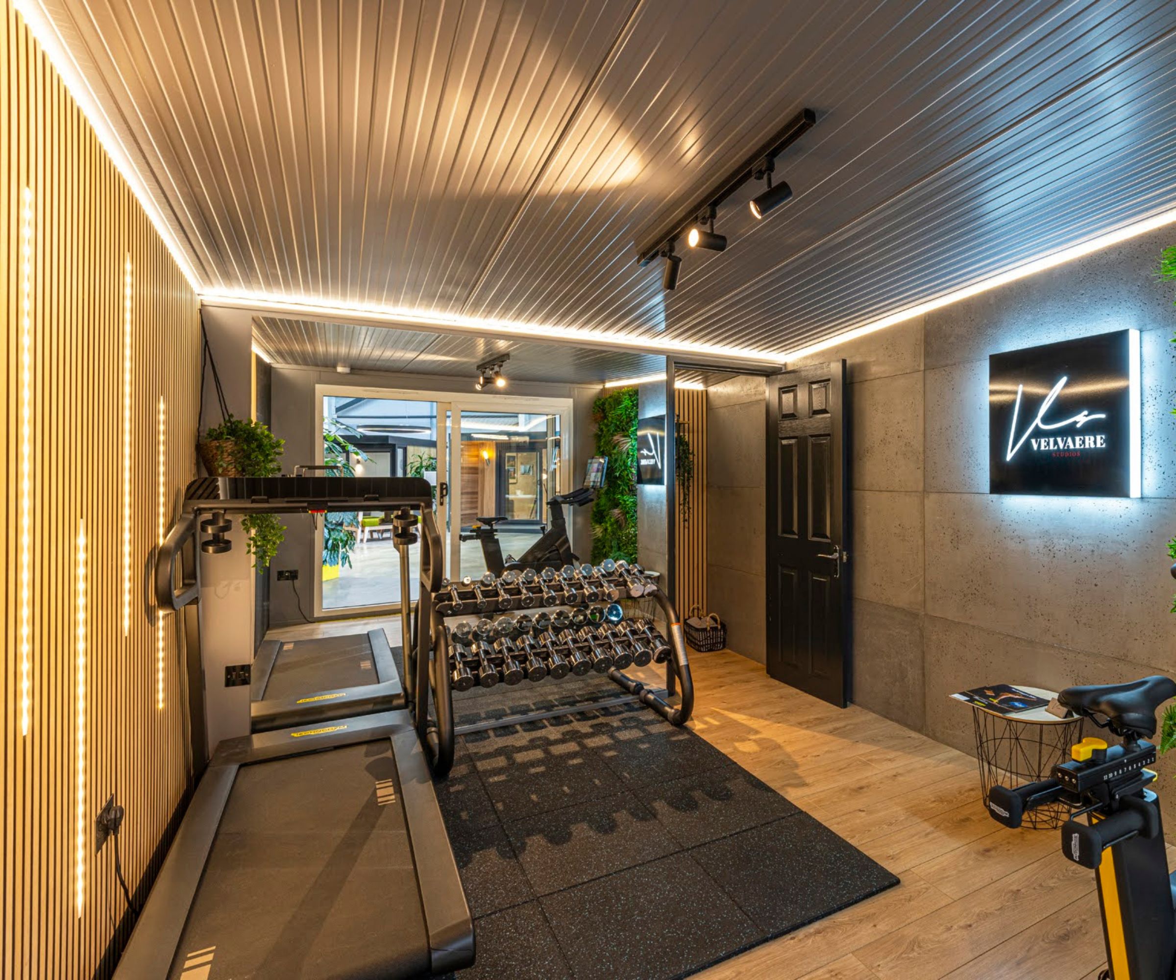 A converted garage home gym with treadmills, weights, and wall-mounted greenery. LED strip lighting runs along the ceiling and walls for a modern look