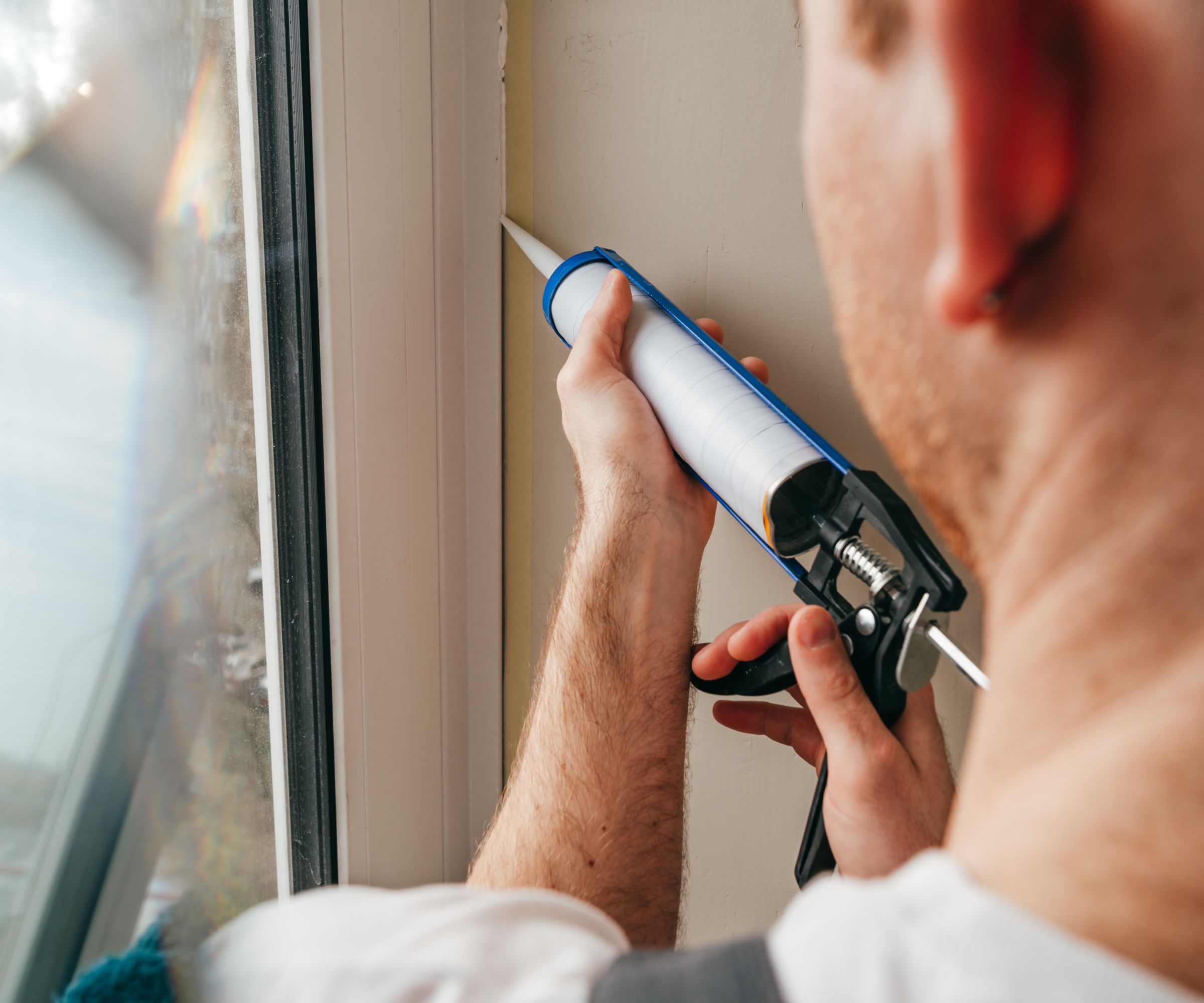 A man wearing overalls sealing cracks between window and trim using waterproof silicone caulk on the balcony.