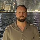 Headshot of a bearded man wearing a light brown shirt in front of water