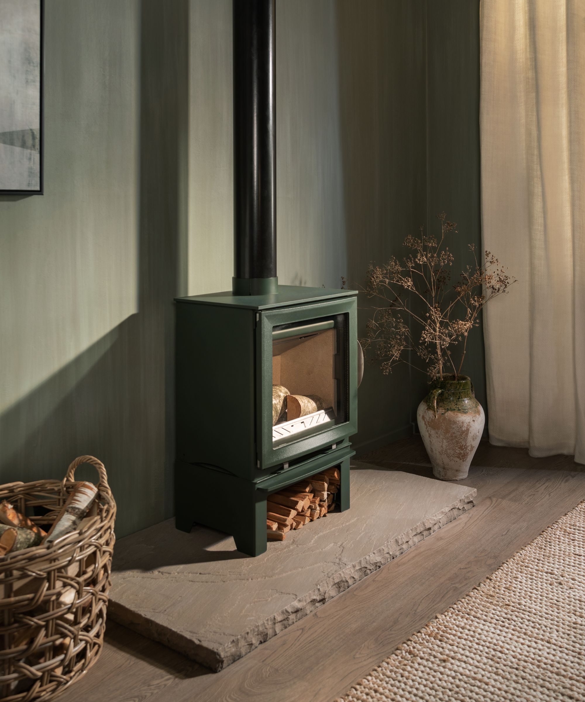 A deep green log burner with a glowing fire sits on a rustic stone hearth. The stove has a small log store beneath, filled with neatly stacked kindling.
