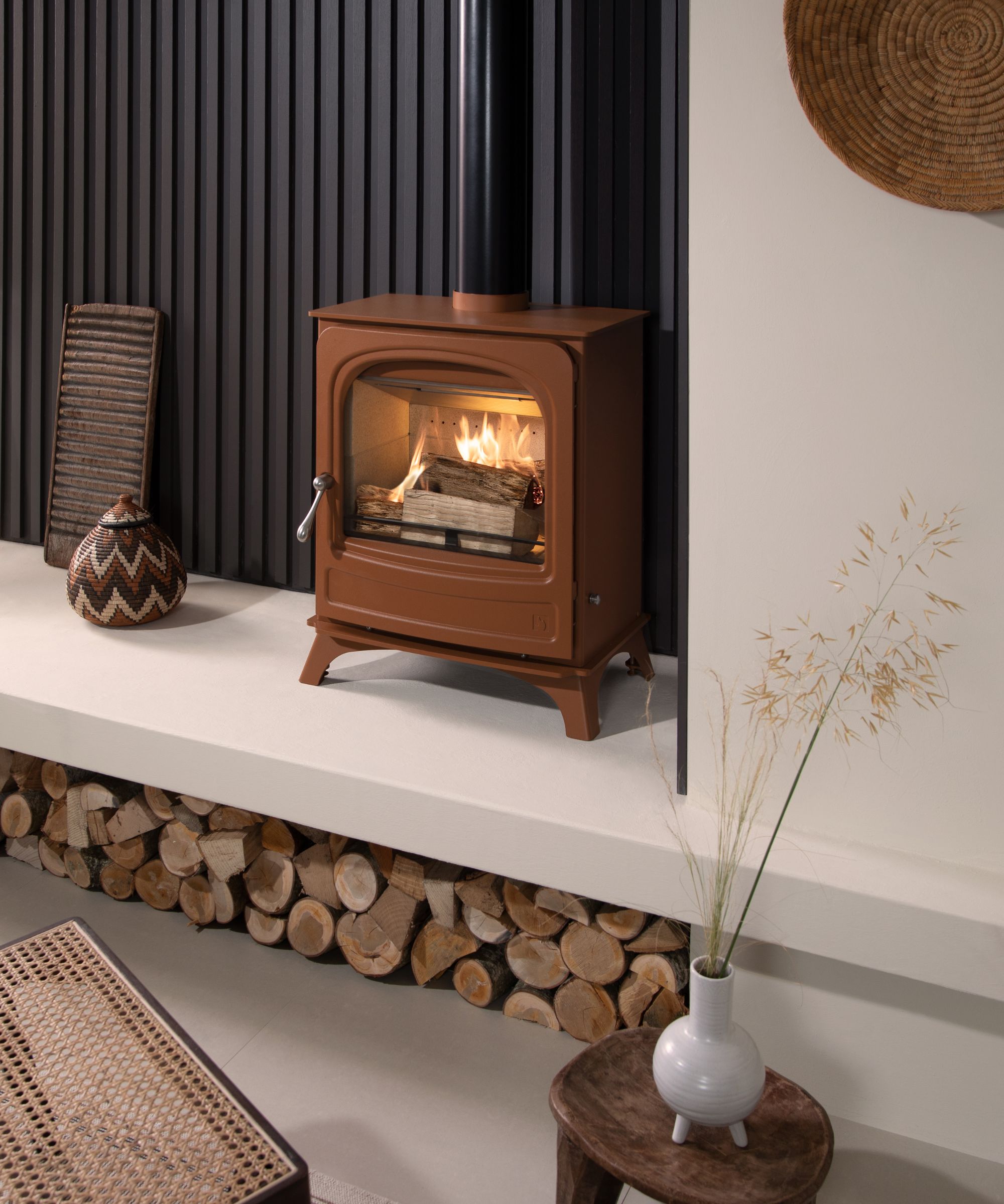 A modern log burner in a warm terracotta colour sits against a wall with a sleek black fluted backdrop. The stove glows with a fire inside, set on a white raised hearth with a log store beneath, neatly stacked with cut firewood.