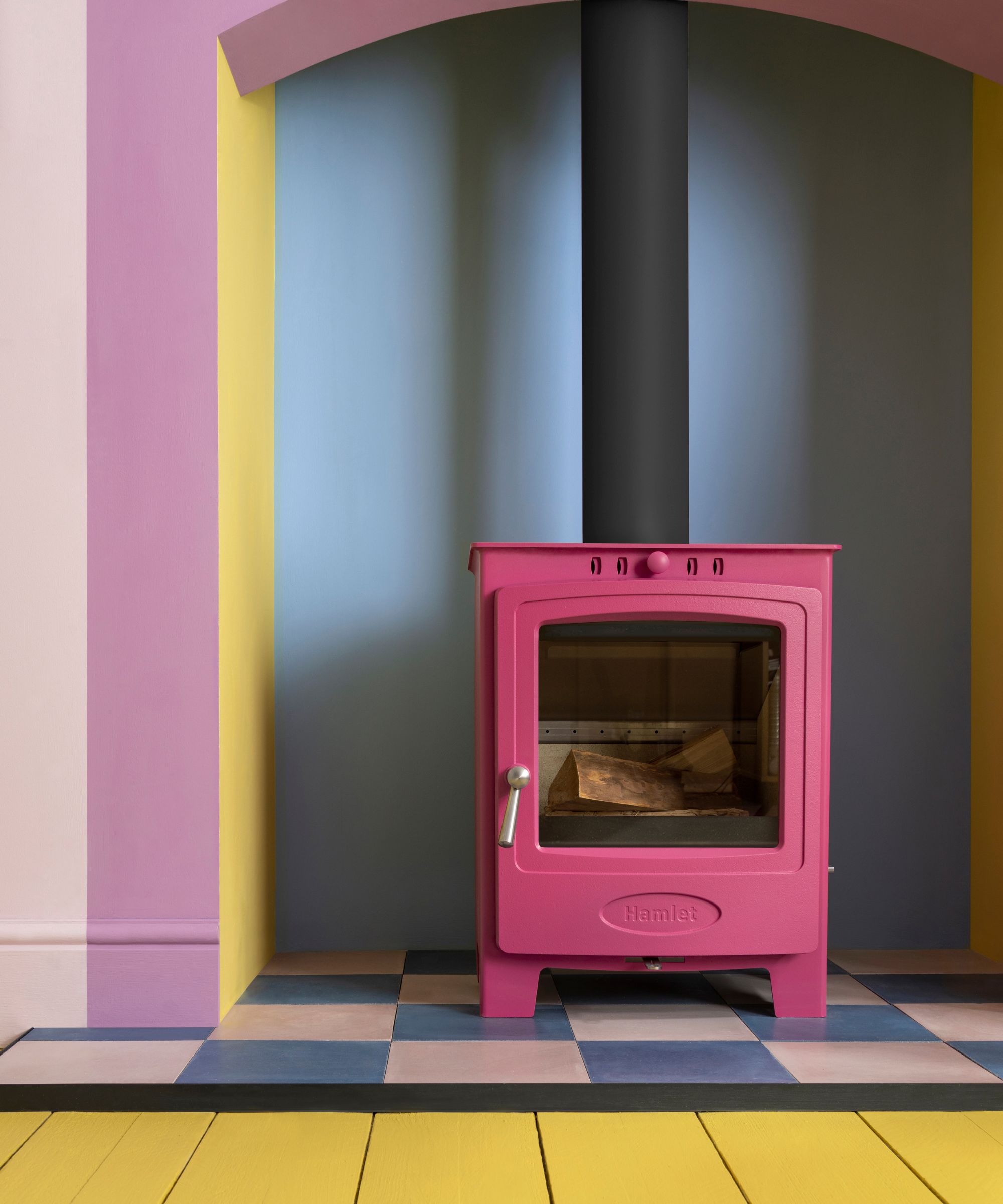 A bold pink log burner stands out in a colourful alcove, framed by walls painted in pastel pink, yellow, and blue. The hearth features a chequered tile design in blue and blush tones, complemented by a bright yellow-painted floorboard surround.