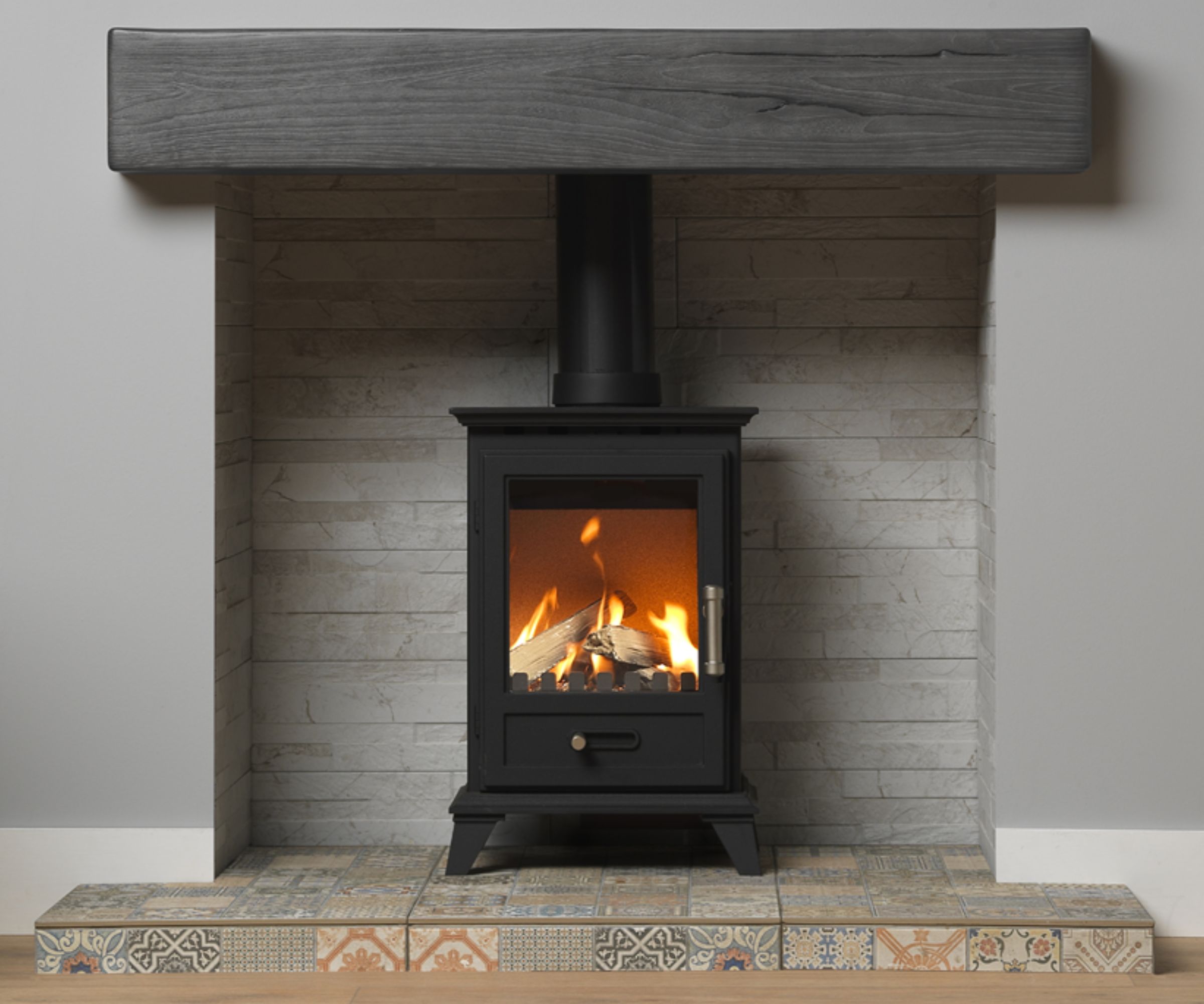 A compact log burner set inside a grey stone-clad fireplace with a dark timber mantel above. The hearth is finished with patterned decorative tiles, adding a colourful and rustic touch.