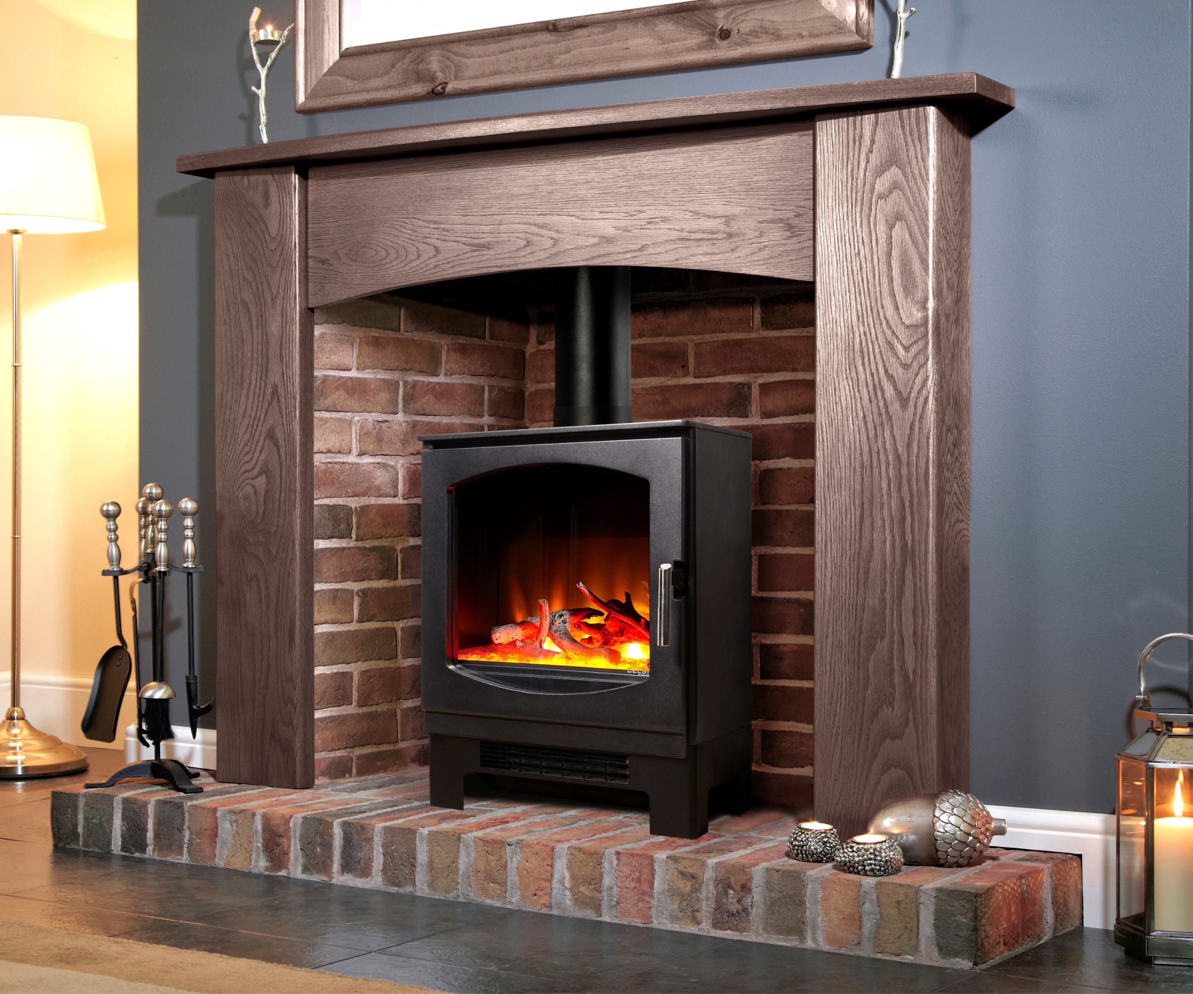 A black log burner with glowing flames sits on a raised brick hearth. The fireplace has a rustic red brick backdrop and a chunky dark wood mantel. A floor lamp, fire tools, and small tealight candles on the hearth add a homely touch.