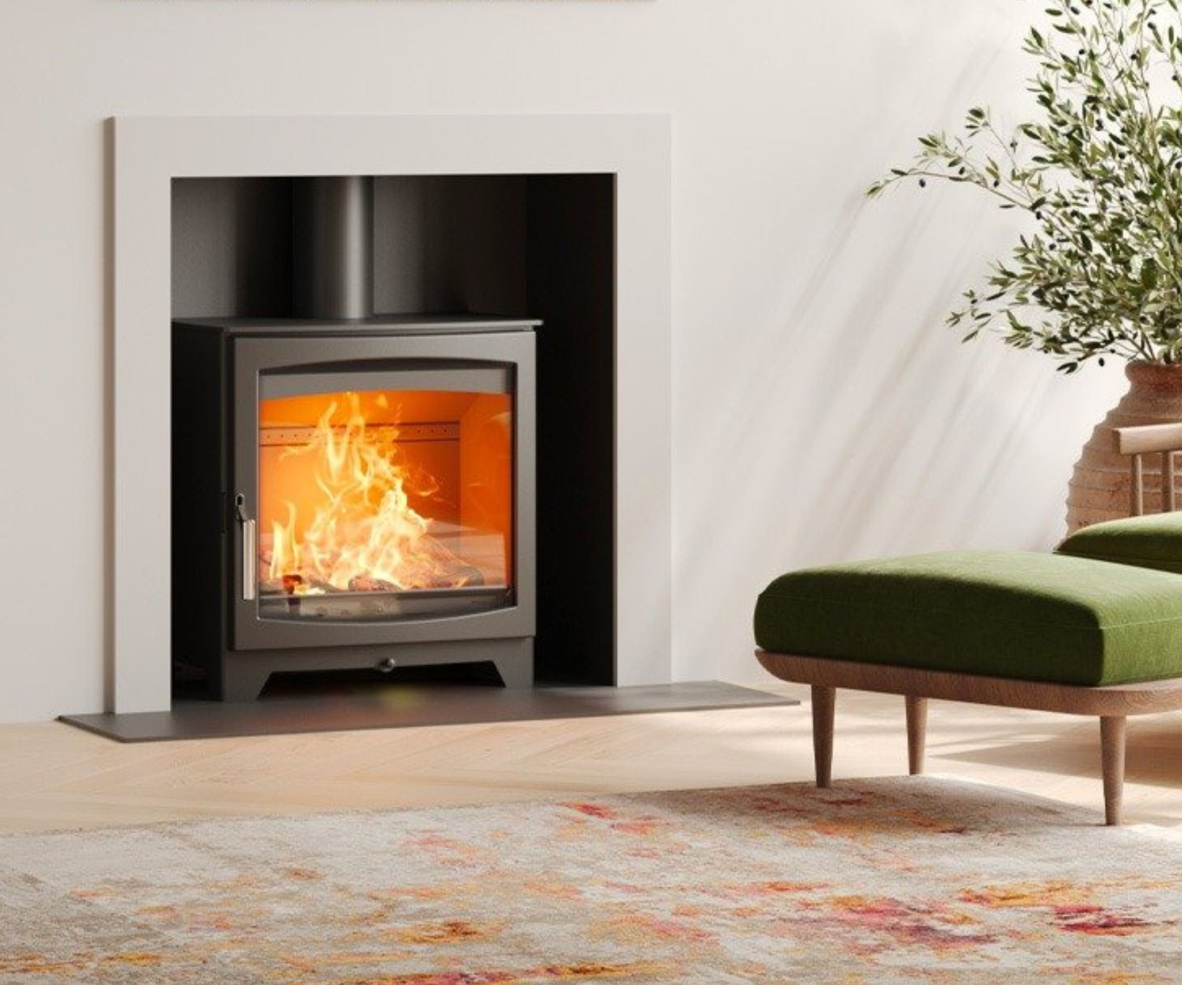 A sleek, modern log burner set into a minimalist white surround. The fire glows brightly, with an olive-green footstool and a rustic vase with foliage adding natural warmth to the light-toned interior.