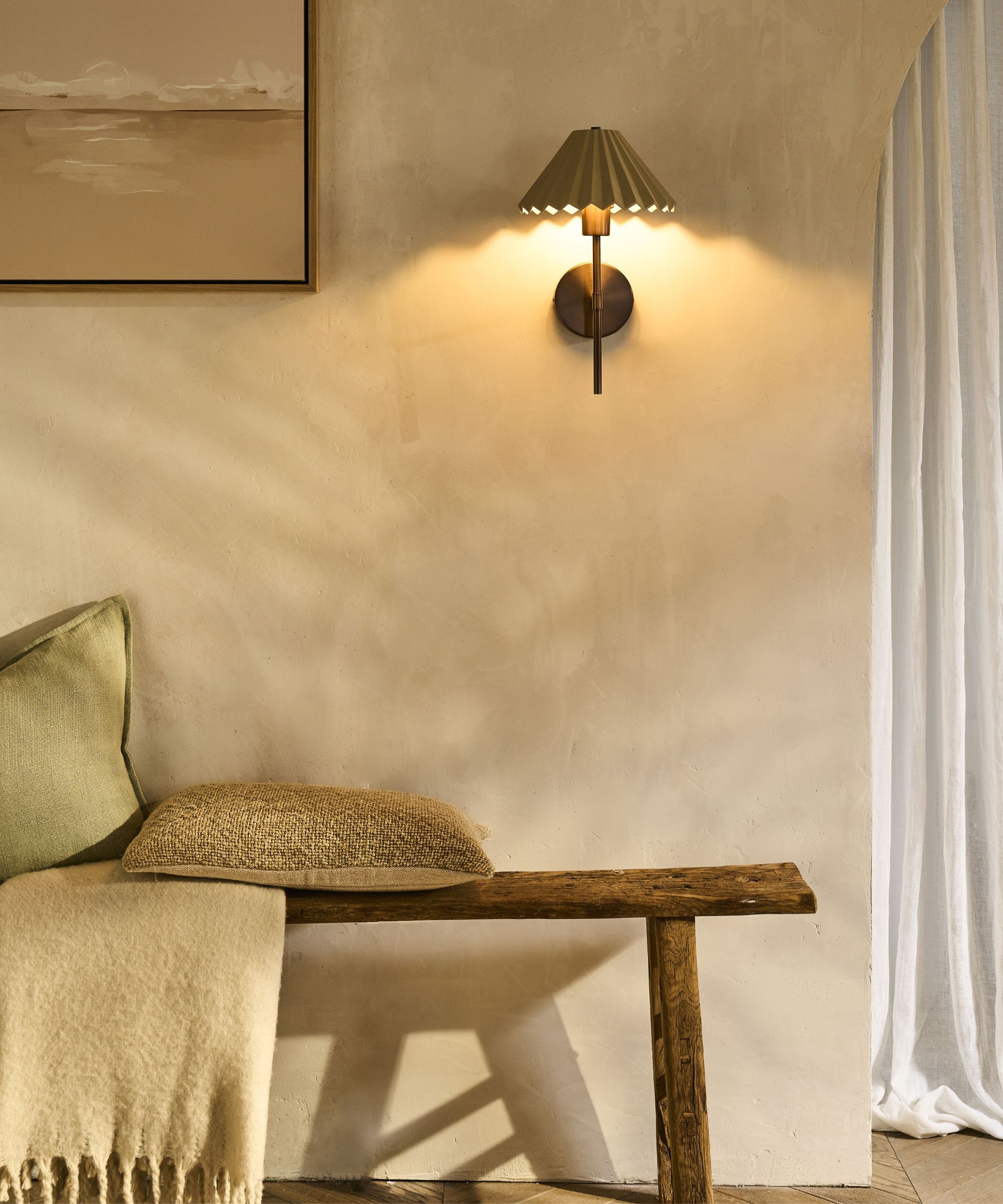 A rustic, softly lit corner featuring a wooden bench with textured cushions and a wool throw. The warm glow of a pleated wall sconce enhances the plastered wall’s natural, tactile finish