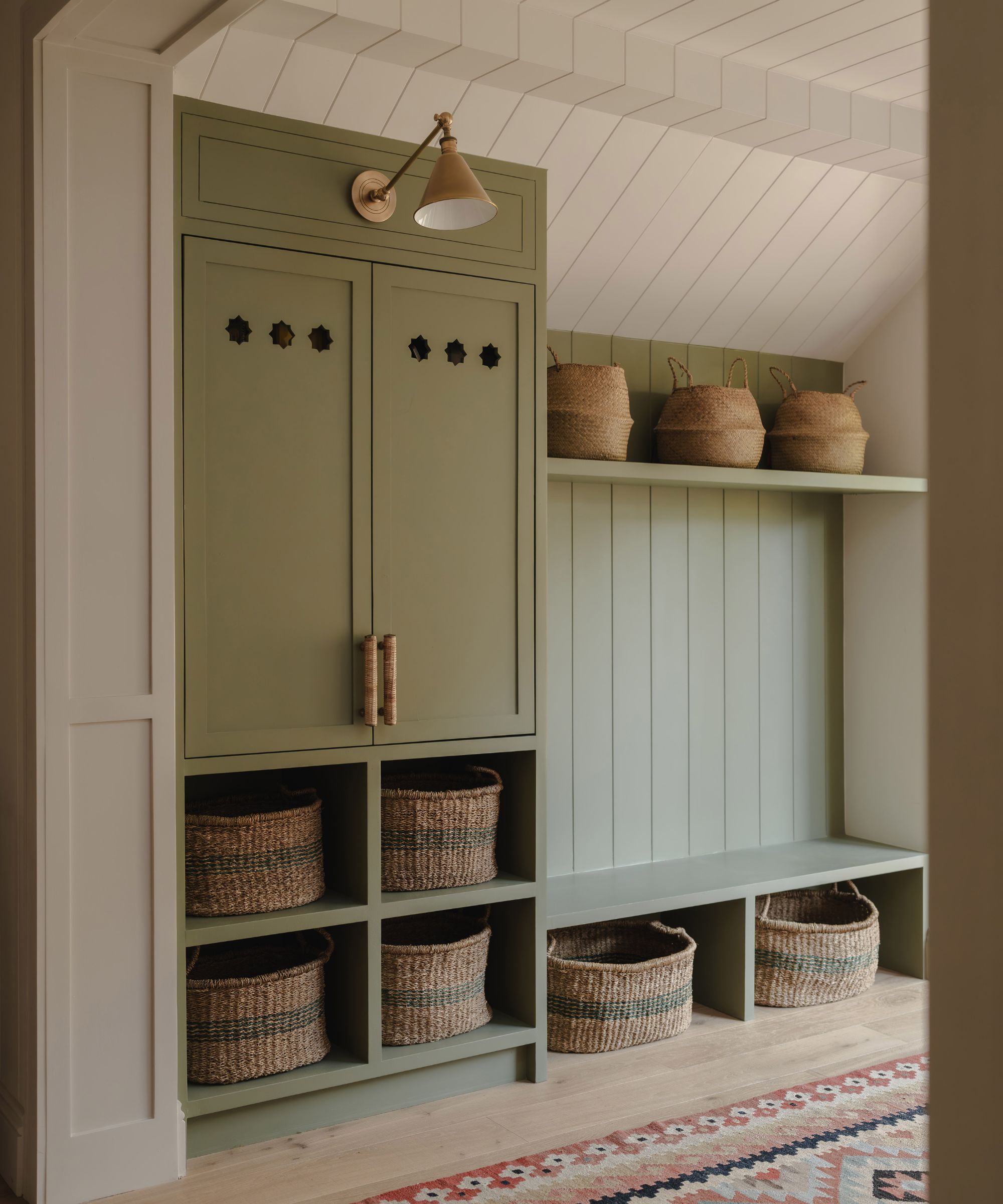 A stylish built-in storage nook painted in soft sage green fills a sloped-ceiling landing. Woven baskets are neatly tucked into cubby holes beneath a wooden bench, with more baskets displayed on a shelf above. A brass wall light adds warmth to the space.