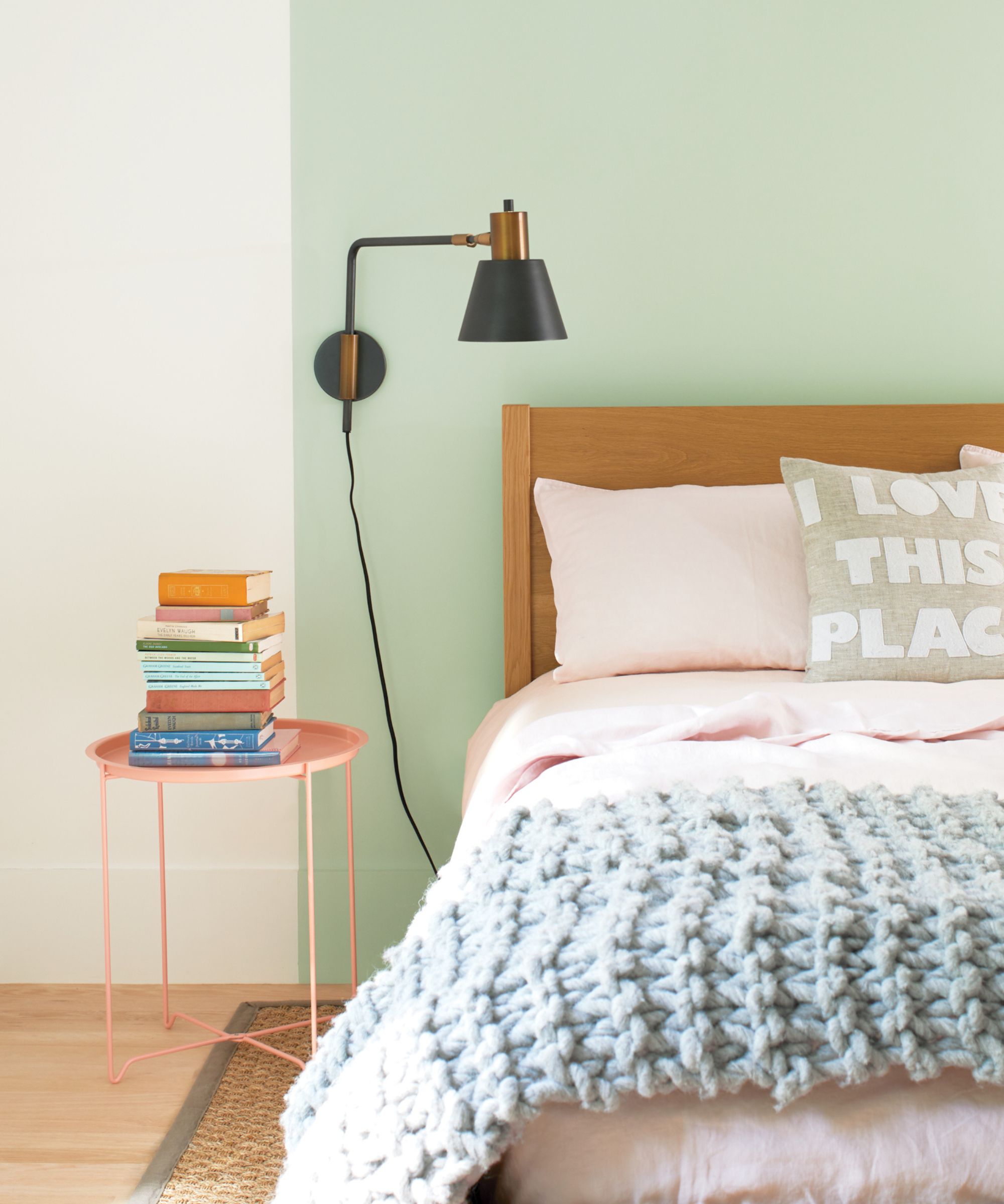 A soft pastel bedroom featuring a mint-green wall, pale pink bedding, and a chunky light blue knitted throw. A black and brass wall light hangs above a pink side table stacked with colourful books.