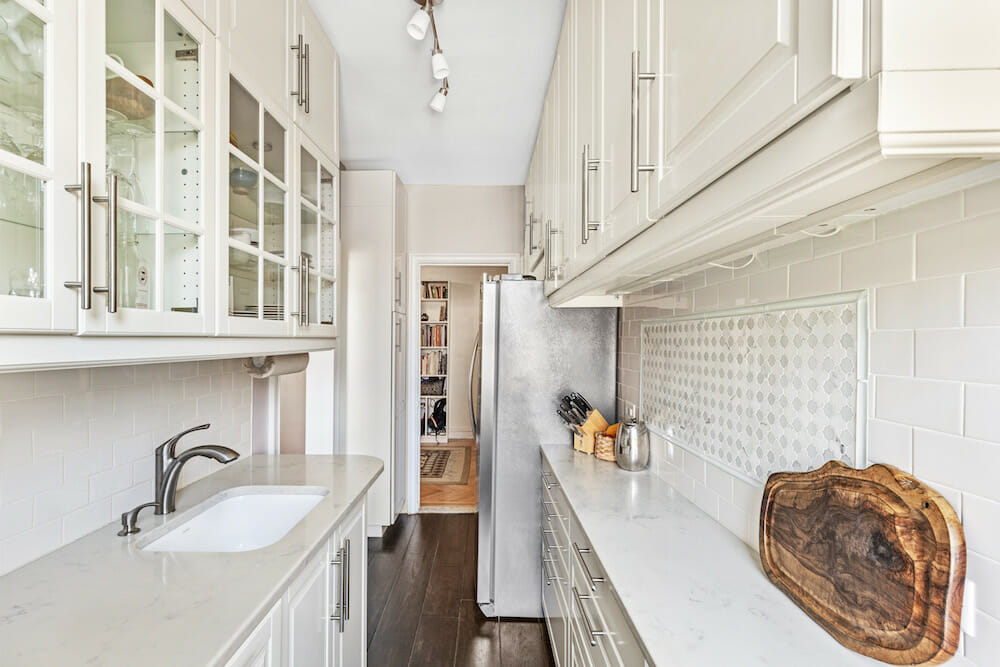 White galley kitchen with glass-front uppers, shallow base cabinets on one side, white quartz counters, subway tile, and a stainless fridge.