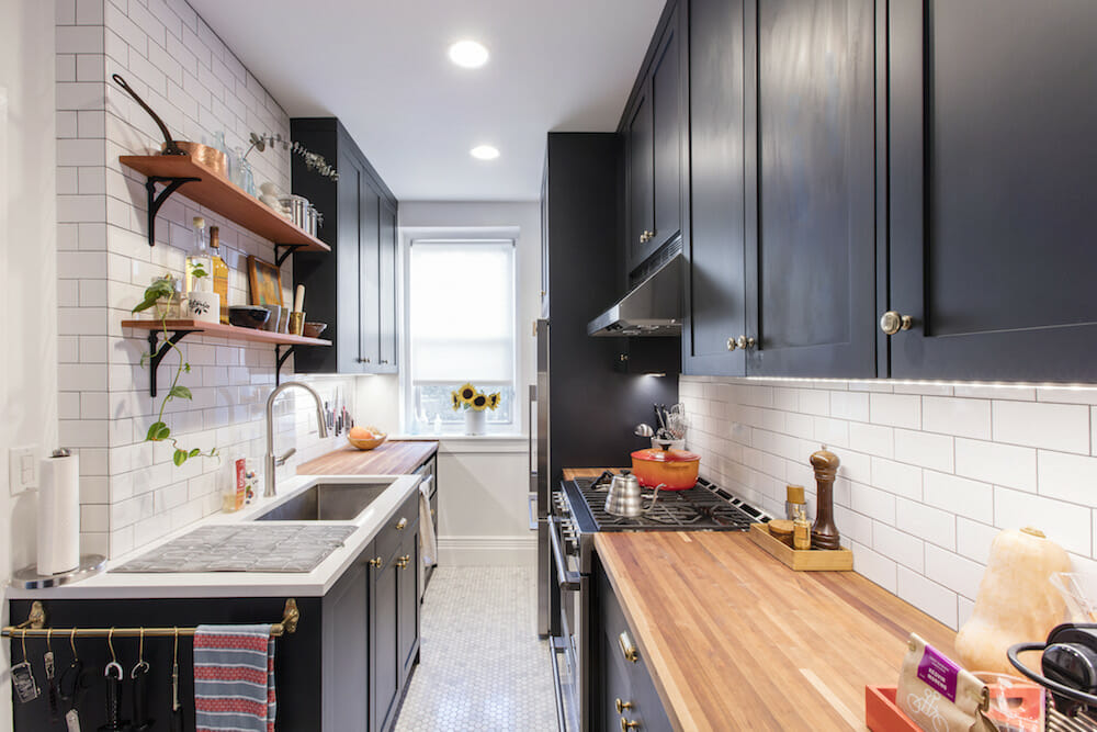 Galley kitchen with shaker cabinets, butcher-block counters, white subway tile, open wood shelves, and a relocated fridge near the window.