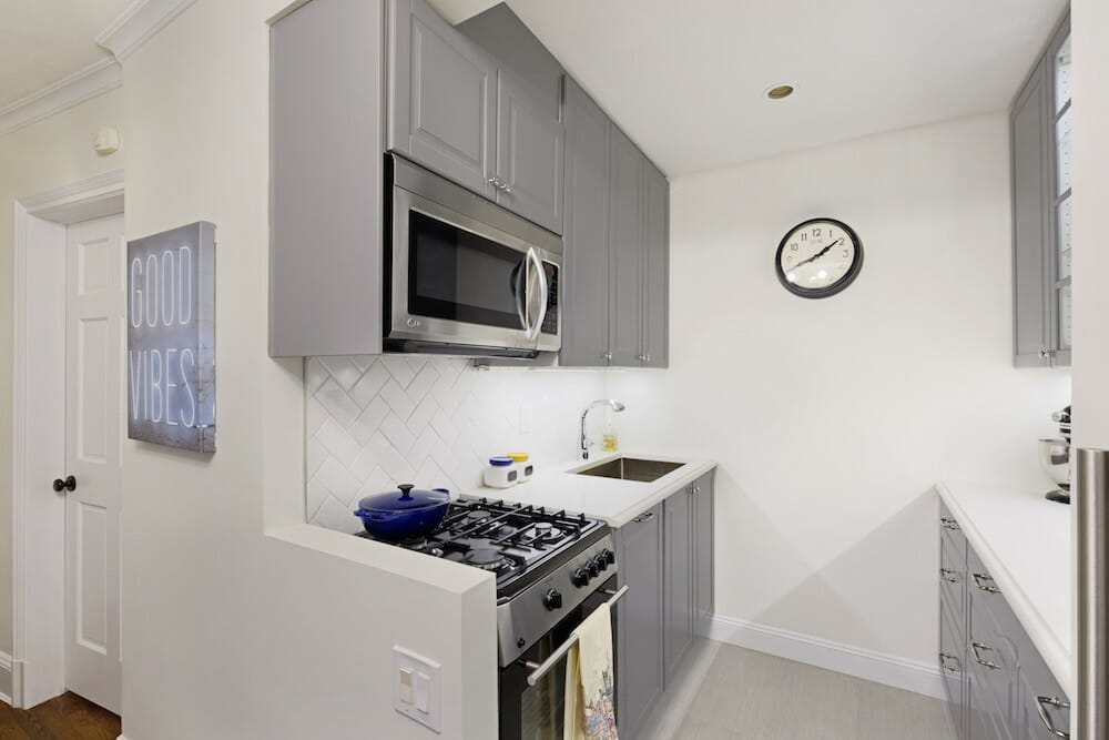 Compact galley with dove gray cabinets, white counters, herringbone subway tile, relocated fridge, gas range, and under-cabinet lighting.