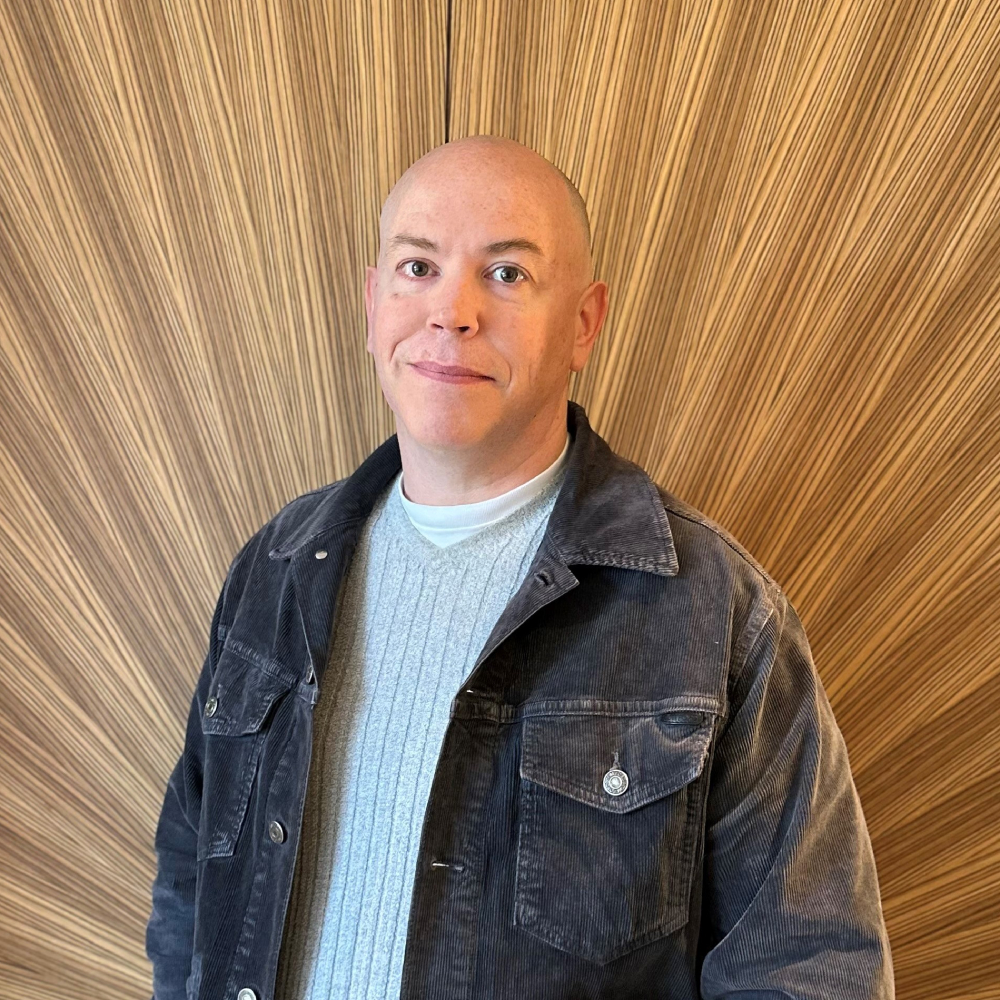 male stood in front of wood panelled wall wearing black denim jacket and grey v neck jumper