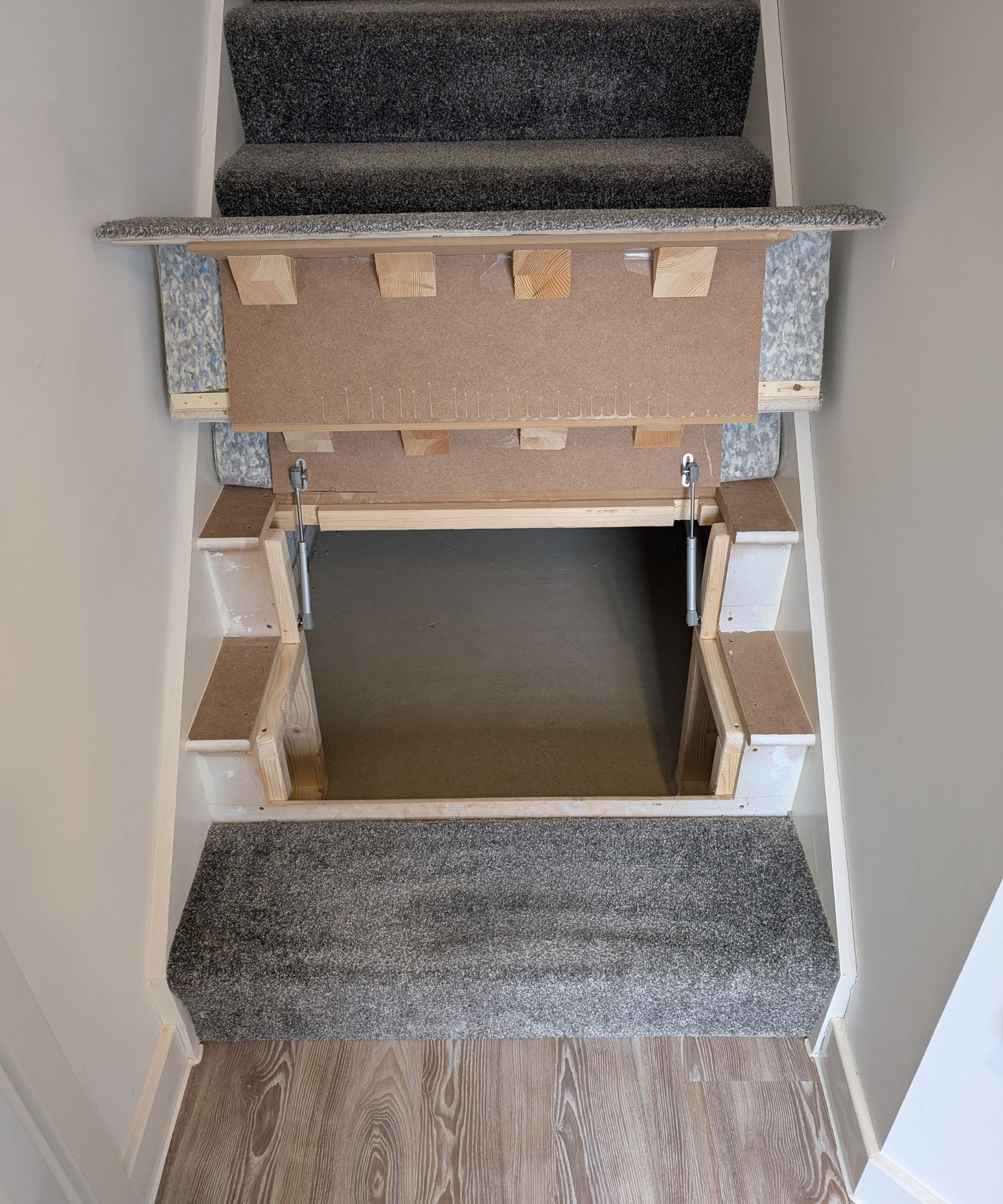 A staircase with grey carpet and pale wood flooring at the base. The lower step lifts open to expose an under-stair storage area, framed neatly in wood and light paintwork.