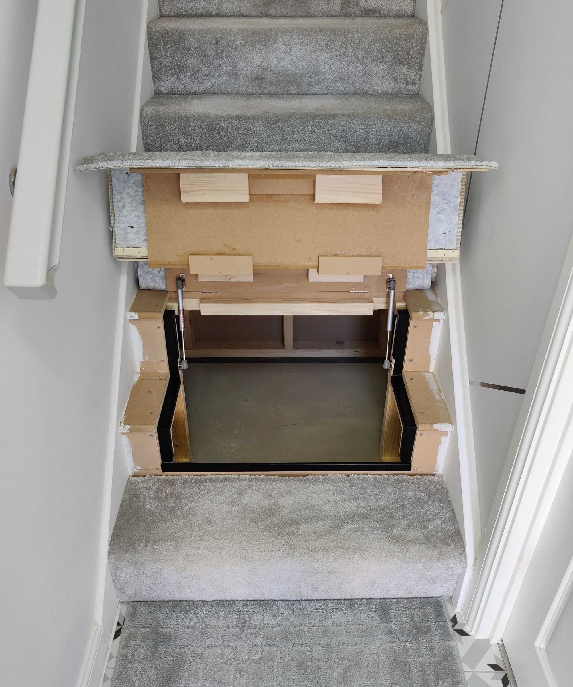 A staircase with one step lifted to reveal a hidden storage compartment beneath. The mechanism is visible, showing wooden supports and metal hinges within a white and grey interior.