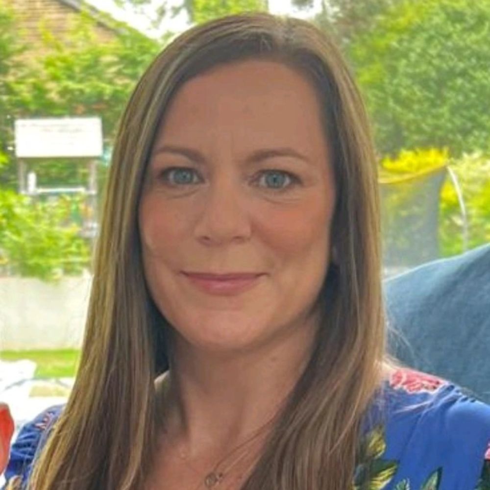 A woman with straight light brown hair and blue eyes smiles gently at the camera. She’s wearing a floral blue top, and the background shows a bright garden scene.