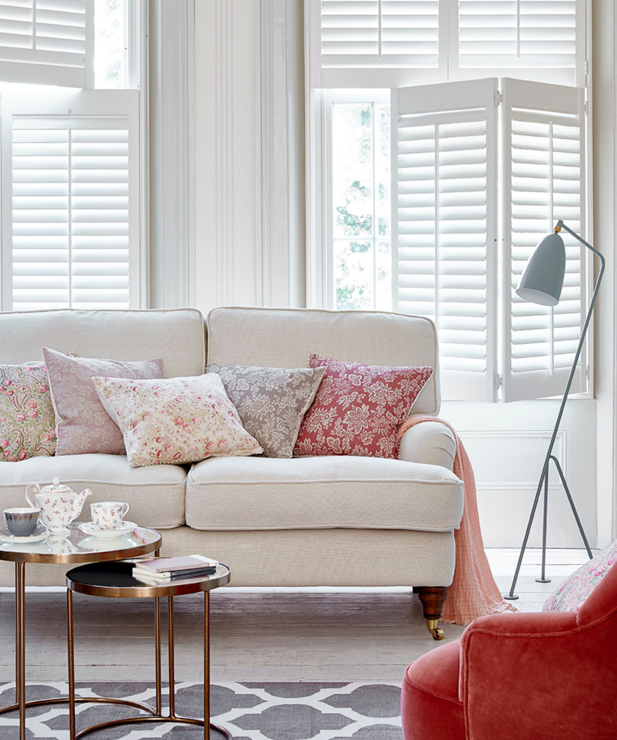 A cosy living room featuring tier-on-tier white shutters. A cream sofa with floral and patterned pink cushions sits alongside a blush armchair and layered coffee tables with tea sets.