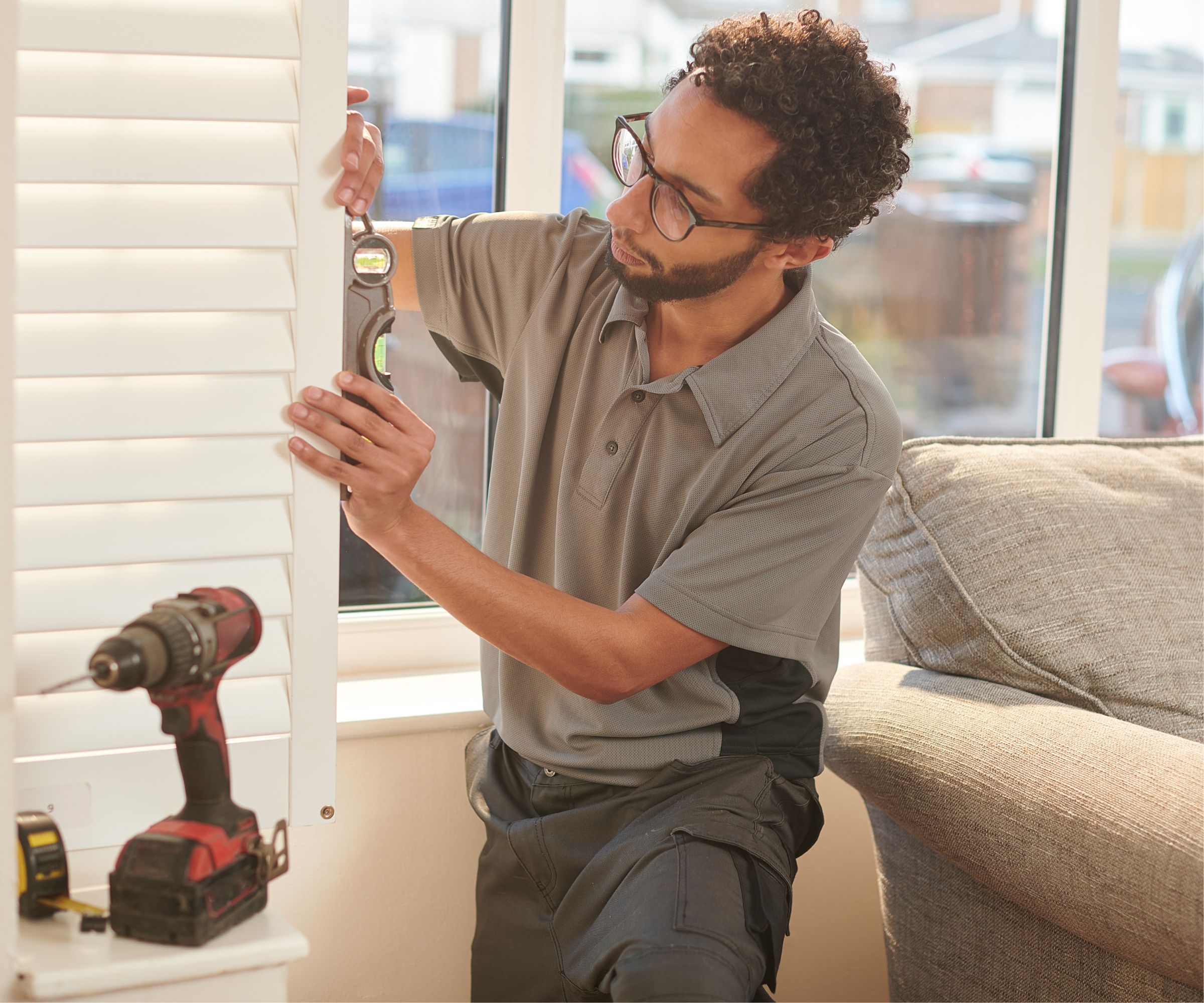 A person carefully installs white plantation shutters beside a living room window, using a spirit level to ensure accuracy. A cordless drill and tape measure sit nearby on the windowsill.