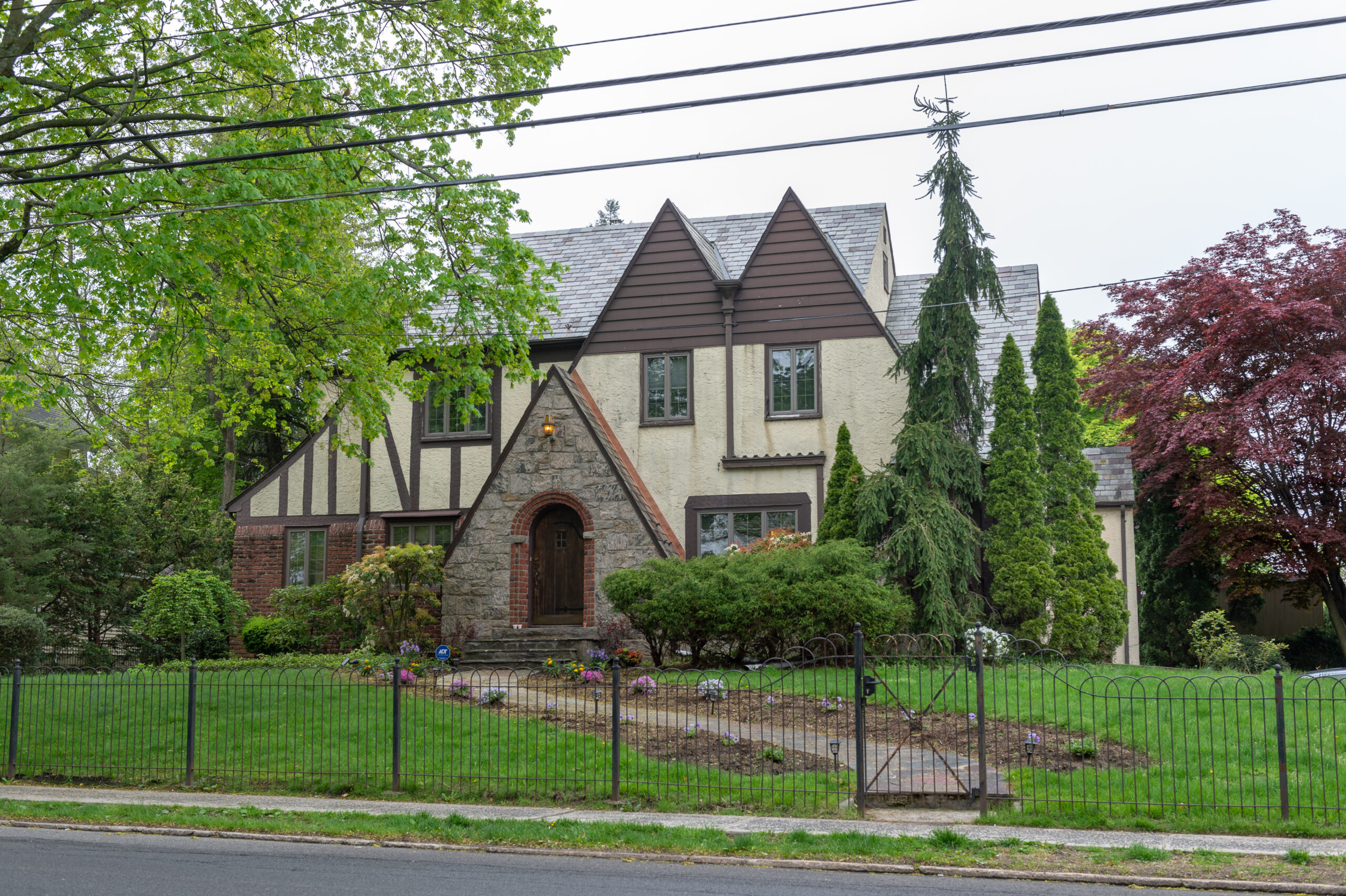 Tudor home exterior with brick, stone entry, stucco, steep rooflines, mature trees, and a front path leading to an arched wood door.