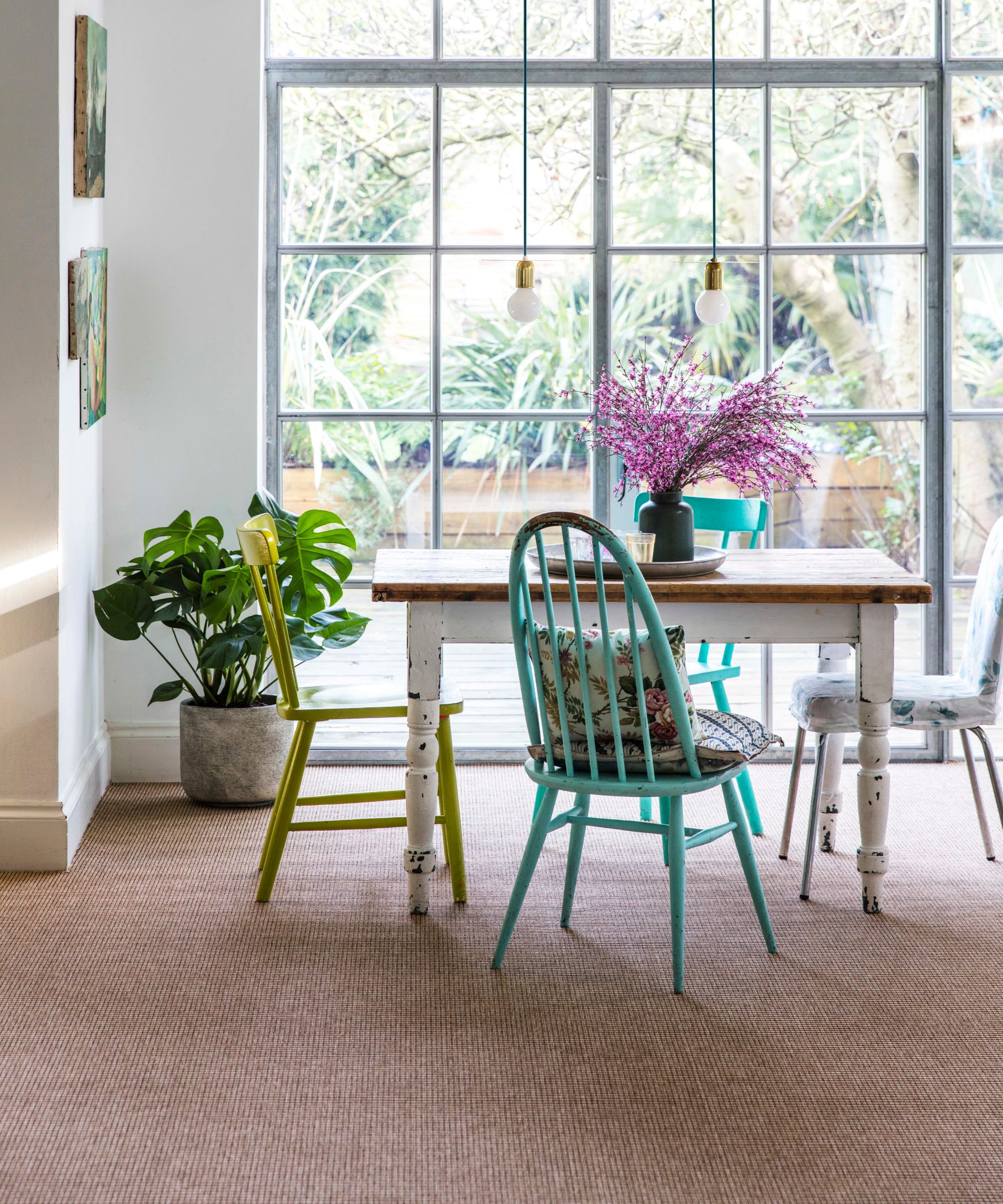 The second image captures a light-filled dining area with floor-to-ceiling windows overlooking a garden. The room features a natural woven carpet under a distressed wooden table paired with colourful, mismatched chairs, creating a cheerful and relaxed atmosphere.