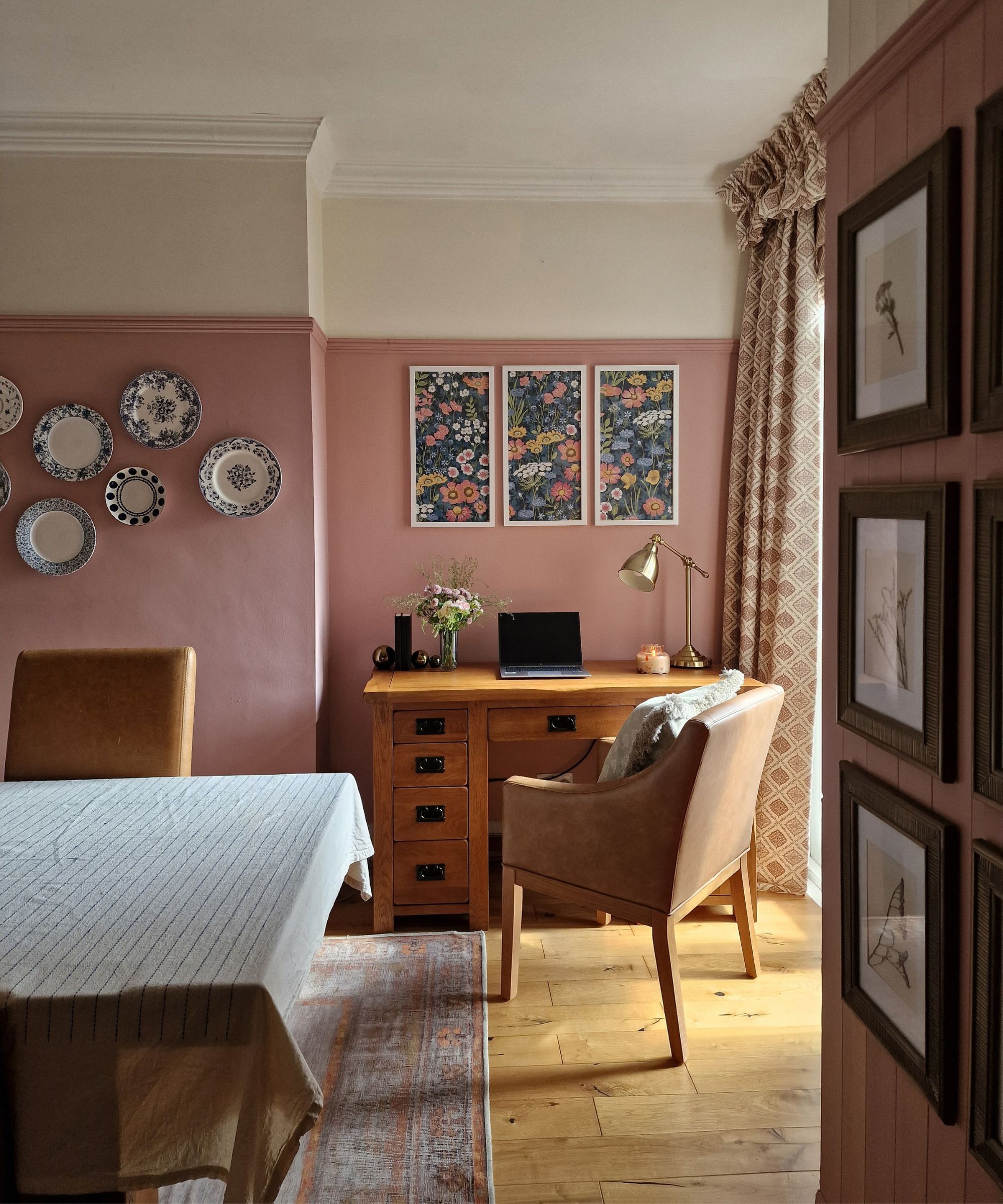 A dining room corner arranged as a compact home office, featuring a wooden desk, leather chair, and laptop. Pink-painted walls, floral artwork and patterned curtains create a warm, traditional feel.