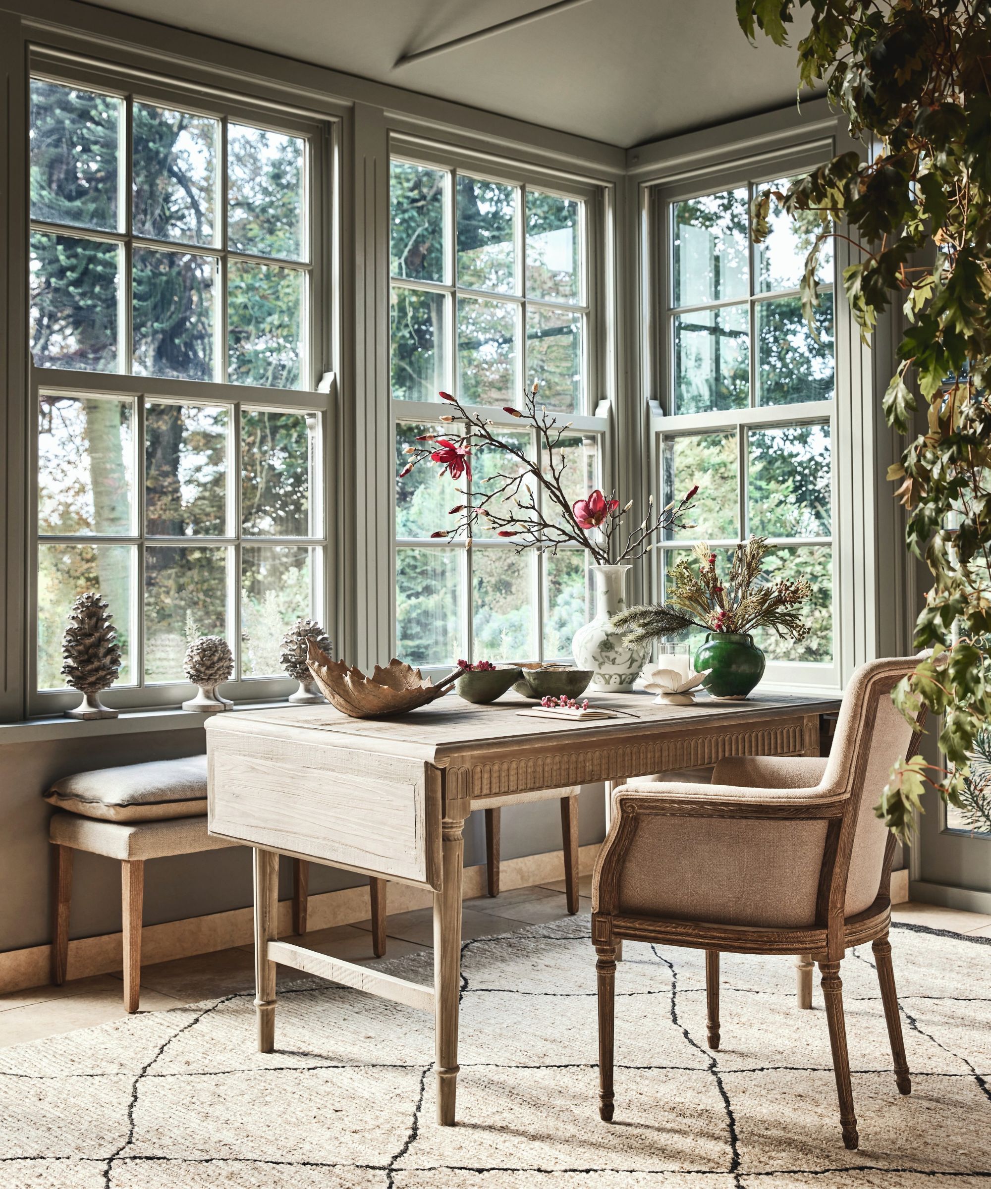 A bright workspace set in a windowed bay, with a pale wooden desk and upholstered chair. Large sash windows frame a garden view, while natural textures and soft, neutral tones enhance the calm atmosphere.