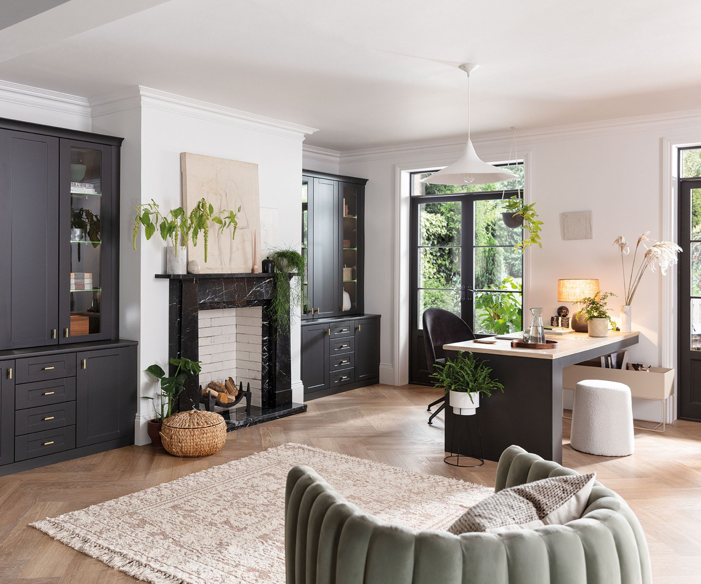 A spacious, light-filled room with dark built-in cabinetry, a marble fireplace and a central desk area. Large windows bring in greenery from outside, while soft furnishings and houseplants add warmth and texture.