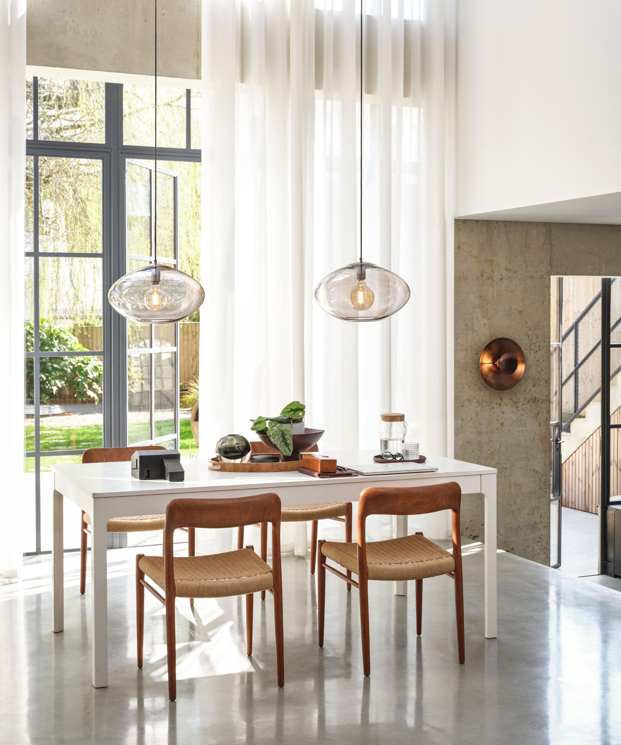 A bright dining space with floor-to-ceiling windows and sheer curtains that soften the daylight. Two glass pendant lights hang above a simple white table surrounded by mid-century style wooden chairs.