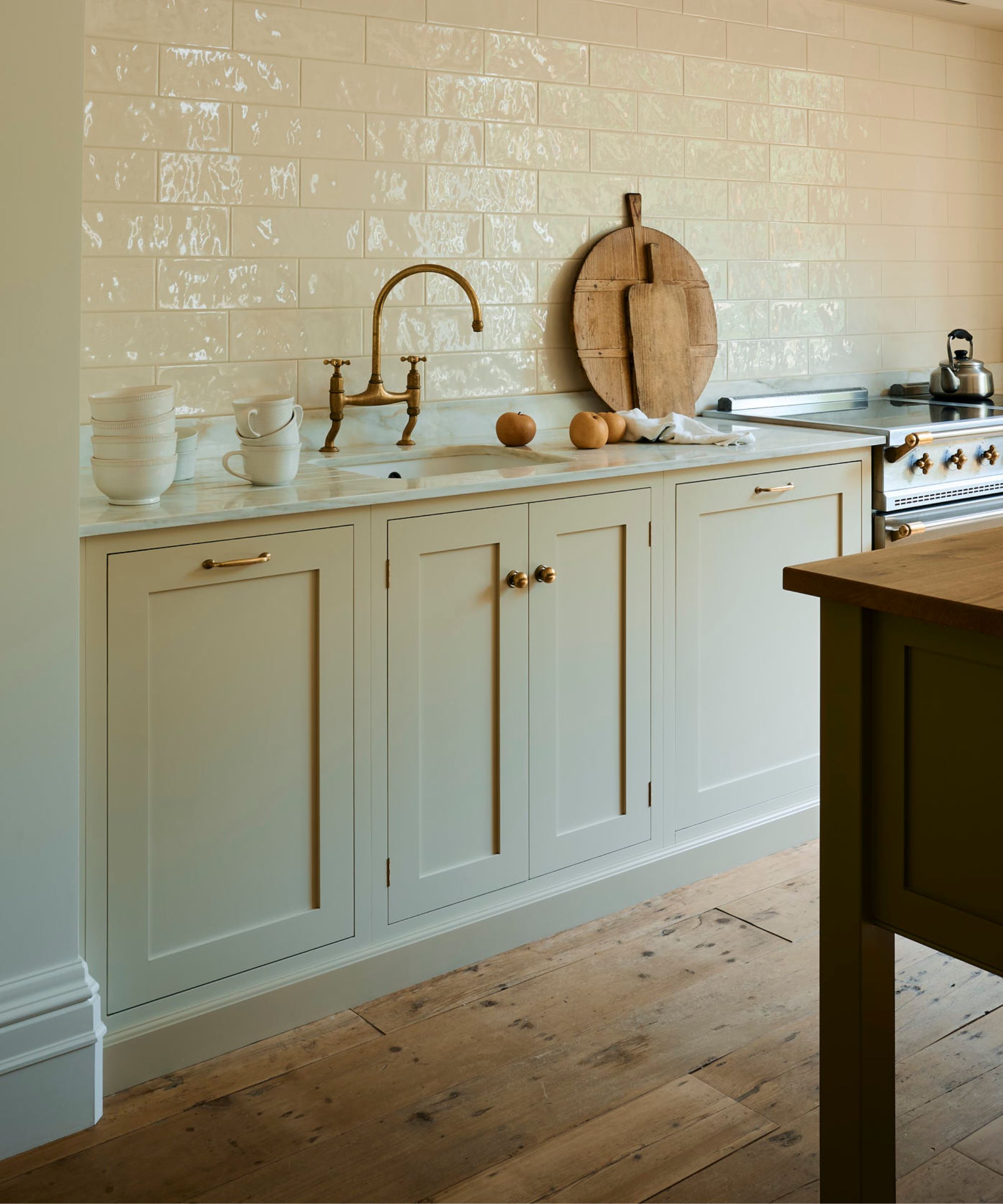 Cream shaker kitchen with brass tap, marble worktop and glossy white tiled splashback, above a wood floor and matching skirting board plinth.