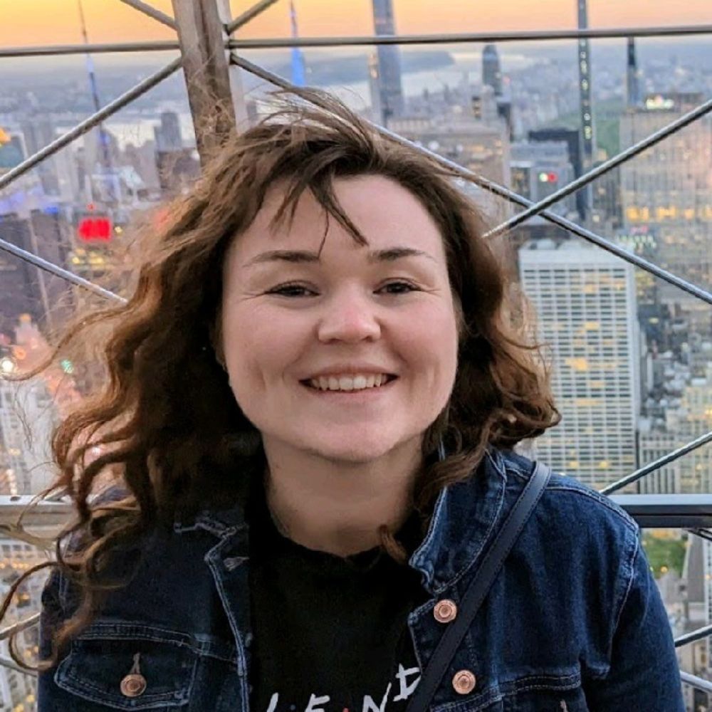 A woman with wavy brown hair smiles warmly at the camera while standing outdoors on a high viewing platform.