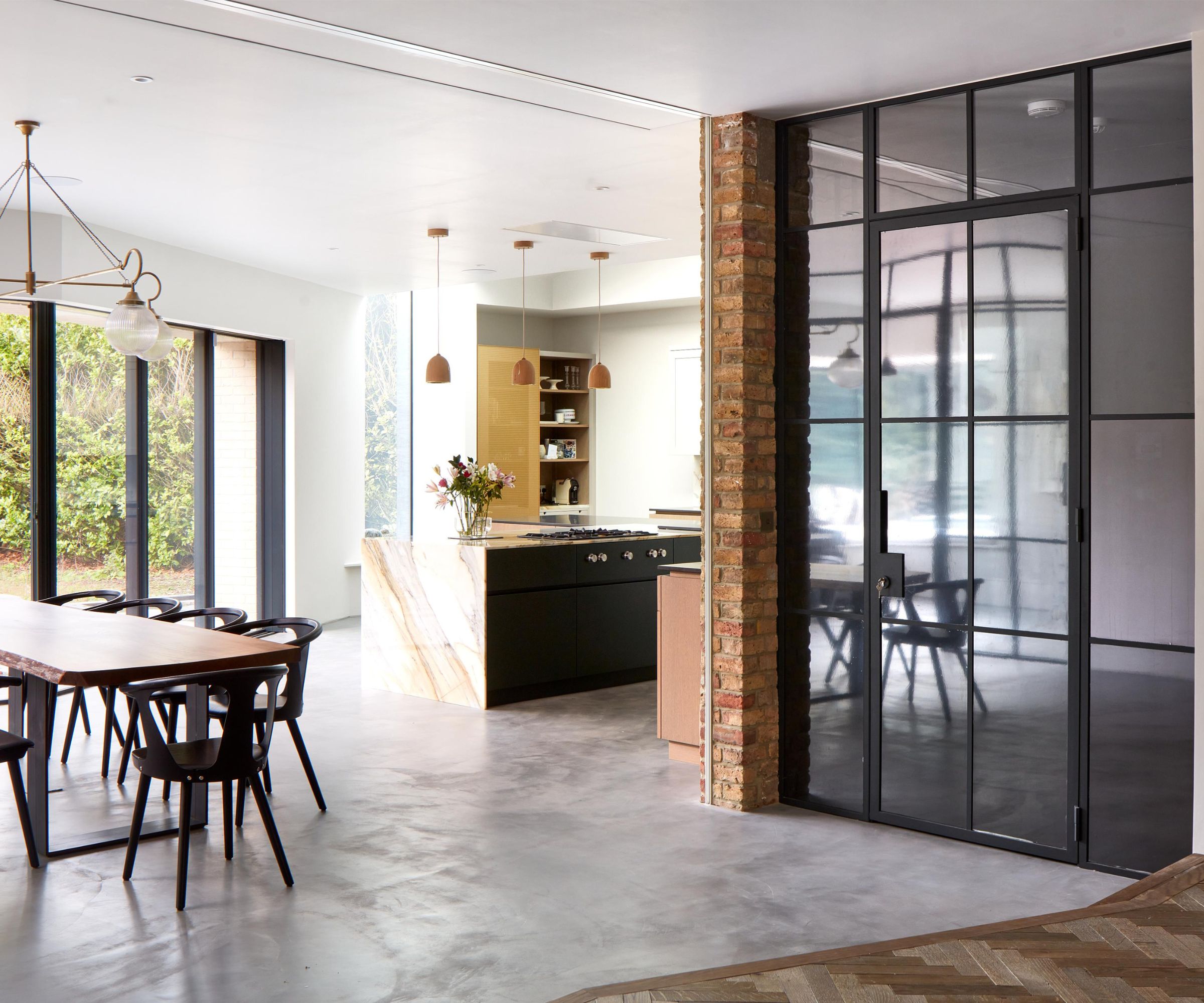Modern open-plan kitchen featuring black framed glass doors enclosing a wine storage room, exposed brick pillar, marble kitchen island and large wooden dining table.