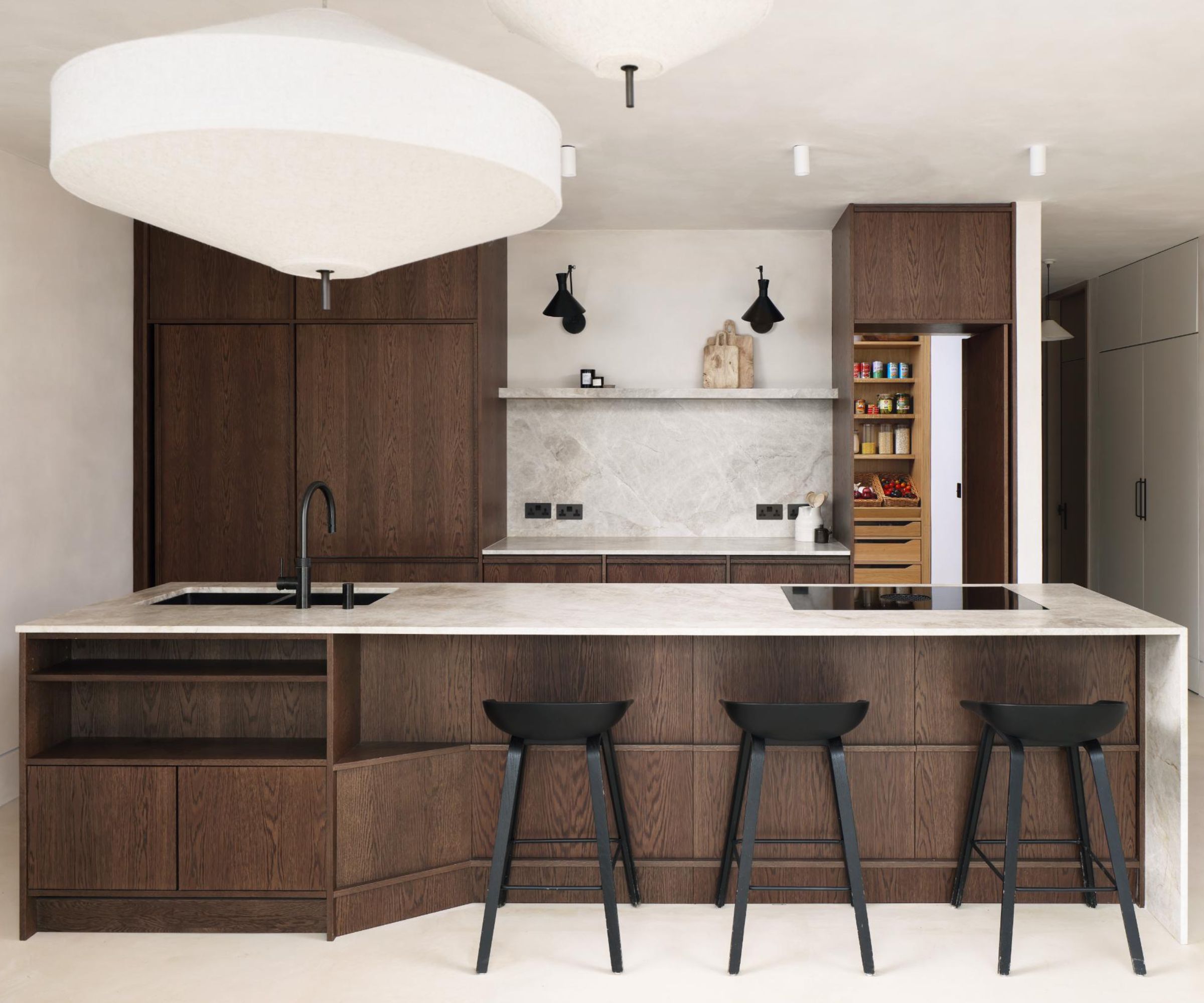 Contemporary kitchen with dark wood cabinetry, a large marble island with black bar stools, and a concealed pantry cupboard opened to reveal neatly organised shelves of food and produce.