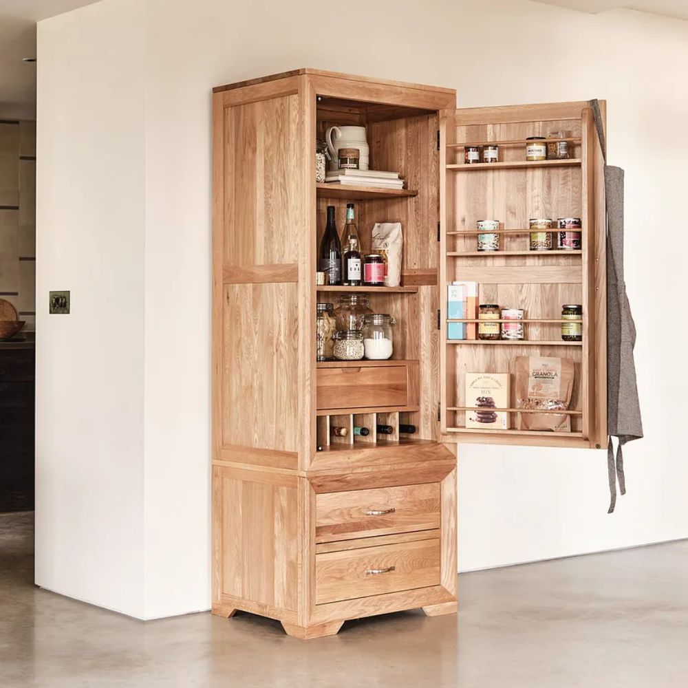 Oak freestanding larder cupboard with open door showing organised shelves of jars, bottles, spices and dry goods, plus built-in wine storage and lower drawers in a modern kitchen.