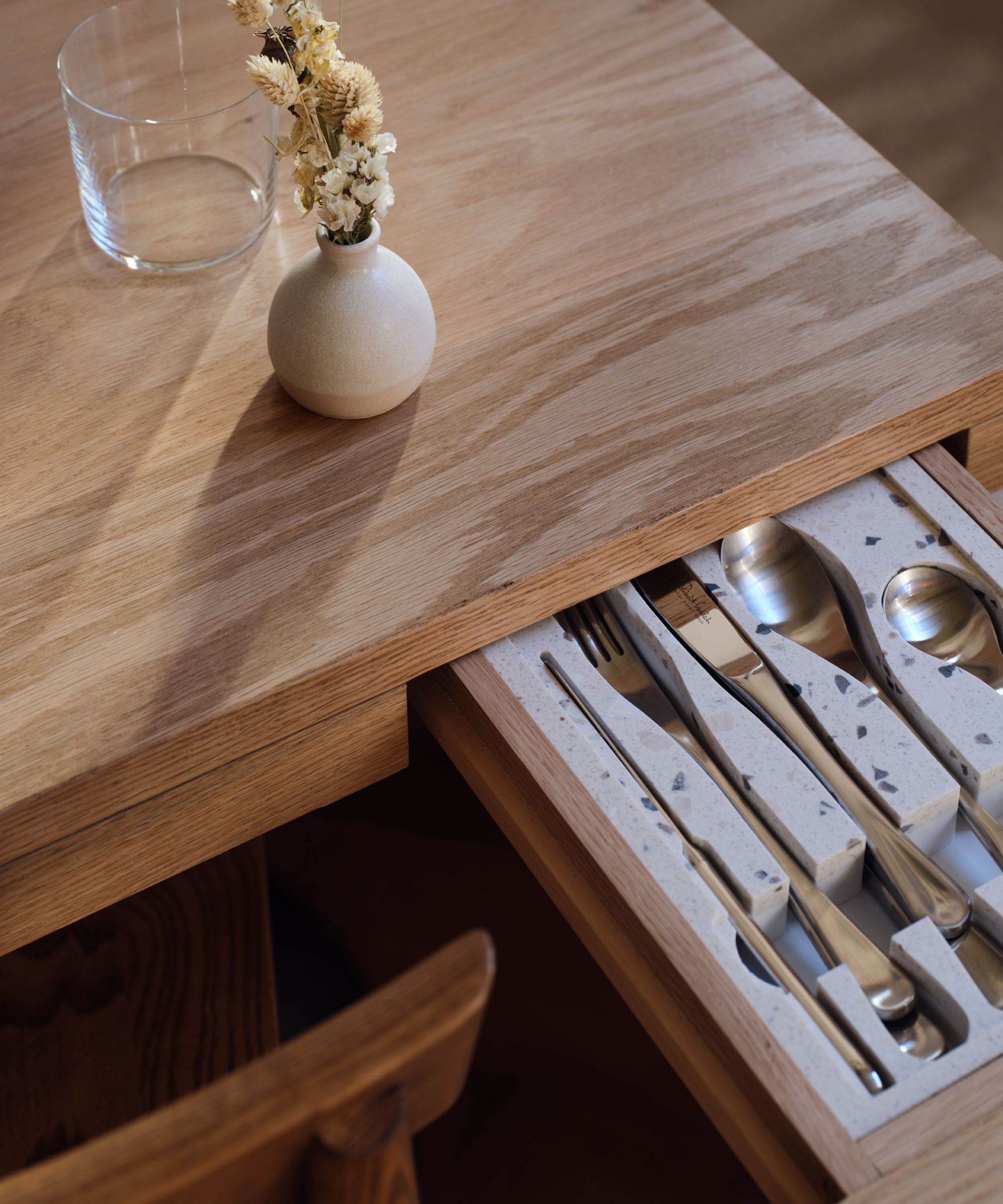 Close-up of a wooden dining table with a concealed cutlery drawer pulled open, featuring terrazzo-style dividers and neatly organised silverware, with a small vase of dried flowers on the tabletop.